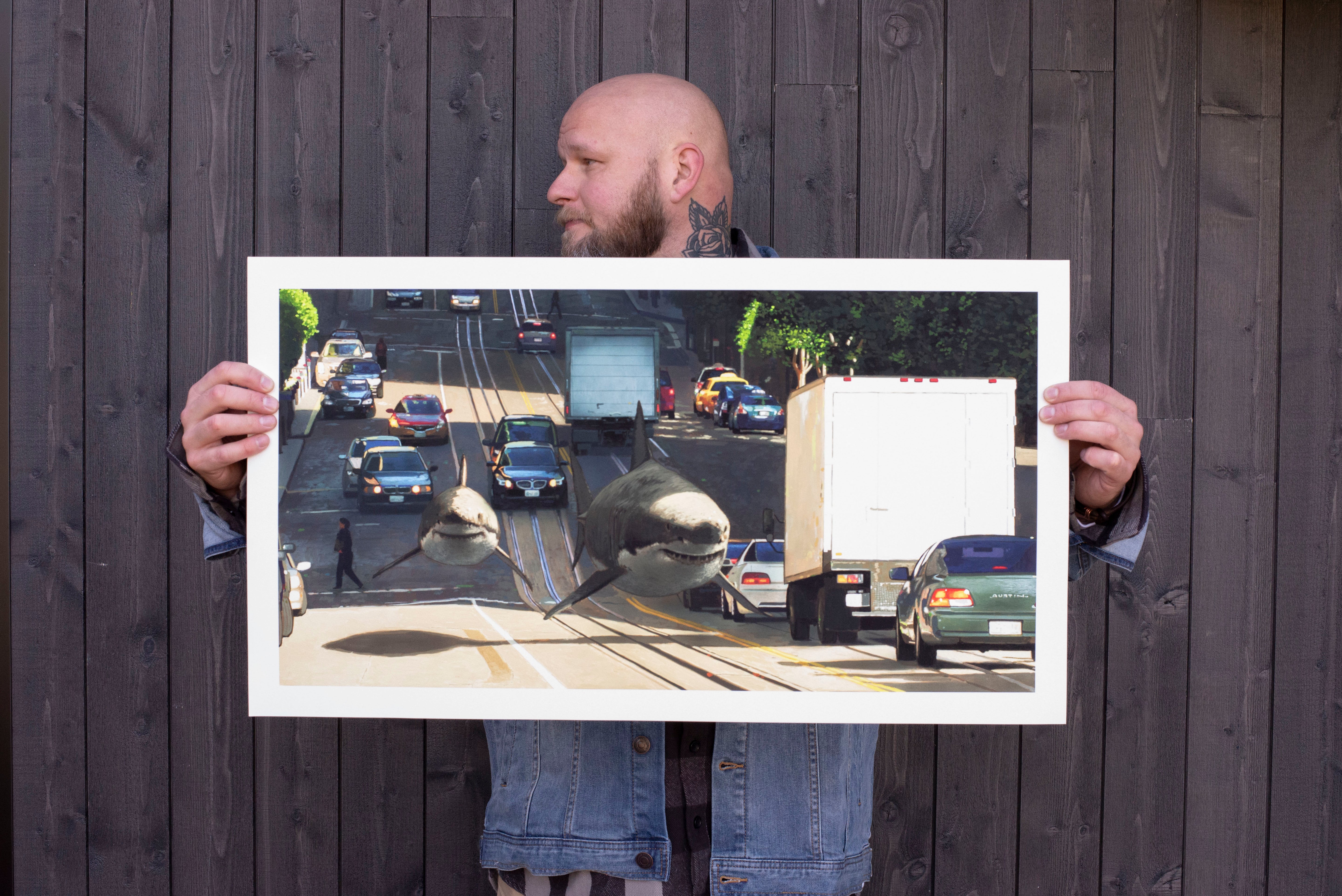 Someone in a denim jacket holds a large, archival pigment print titled Thriving Cities by Chris Austin, illustrating two massive whales swimming down a bustling city street filled with cars and pedestrians, set against a wooden wall.