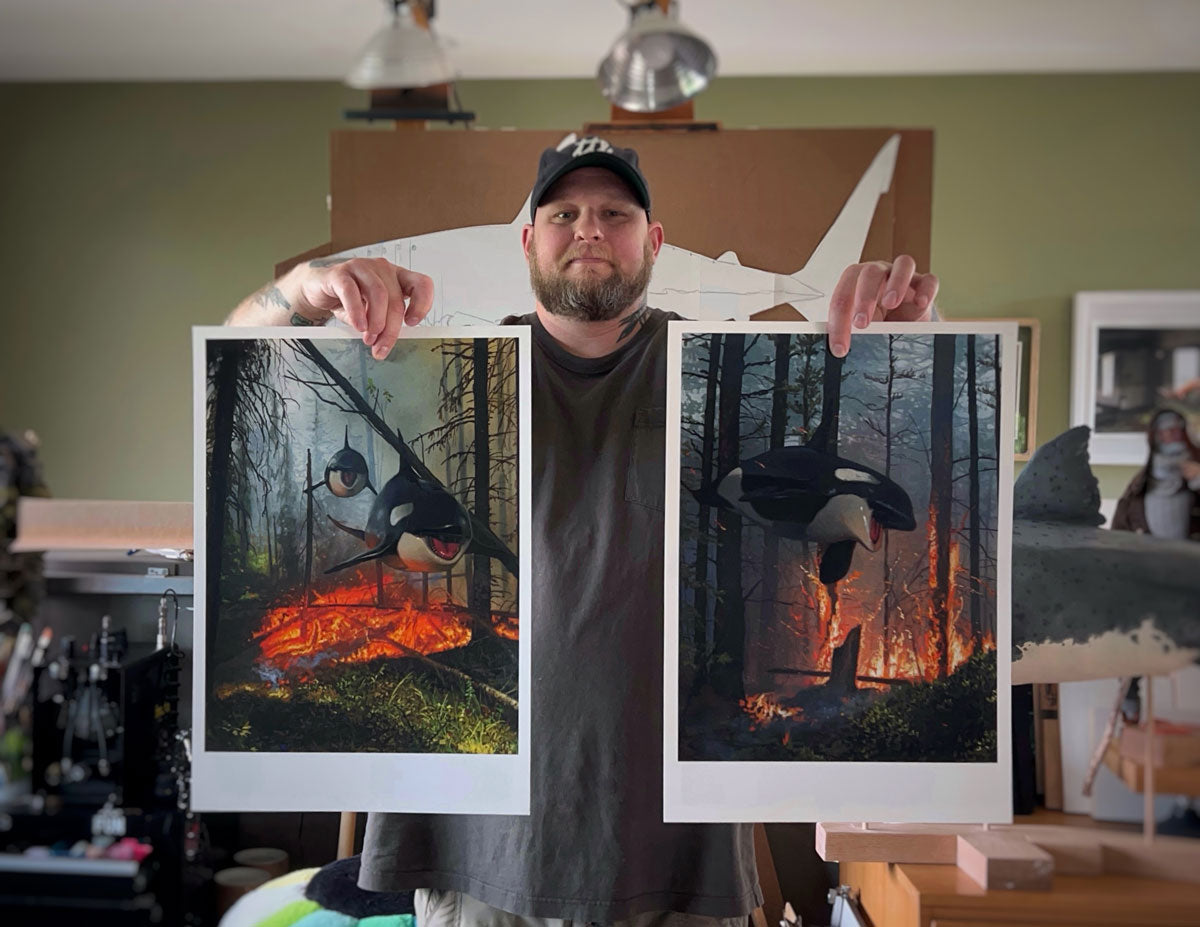 A man in a cap holds Chris Austin’s “Dream and Lost” diptych—paintings of orcas swimming through a forest above wildfires, with distinct lighting and details. Art supplies, a shark model, and signed, numbered prints appear in the background.