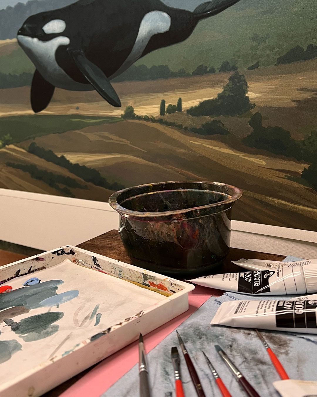 A close-up of an artist’s workspace with paintbrushes, paint tubes, a palette, and "Get On With It"—an orca landscape artwork with hand-painted details by Chris Austin.