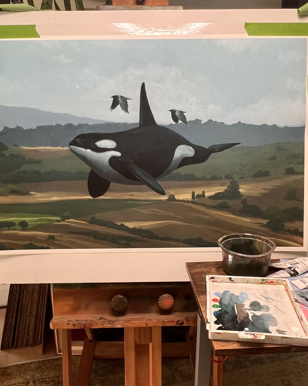Get On With It" by Chris Austin is an archival pigment print with hand painted embellishments, depicting an orca swimming above a rural landscape with hills, trees, and birds. Displayed on an easel with painting supplies in the foreground.