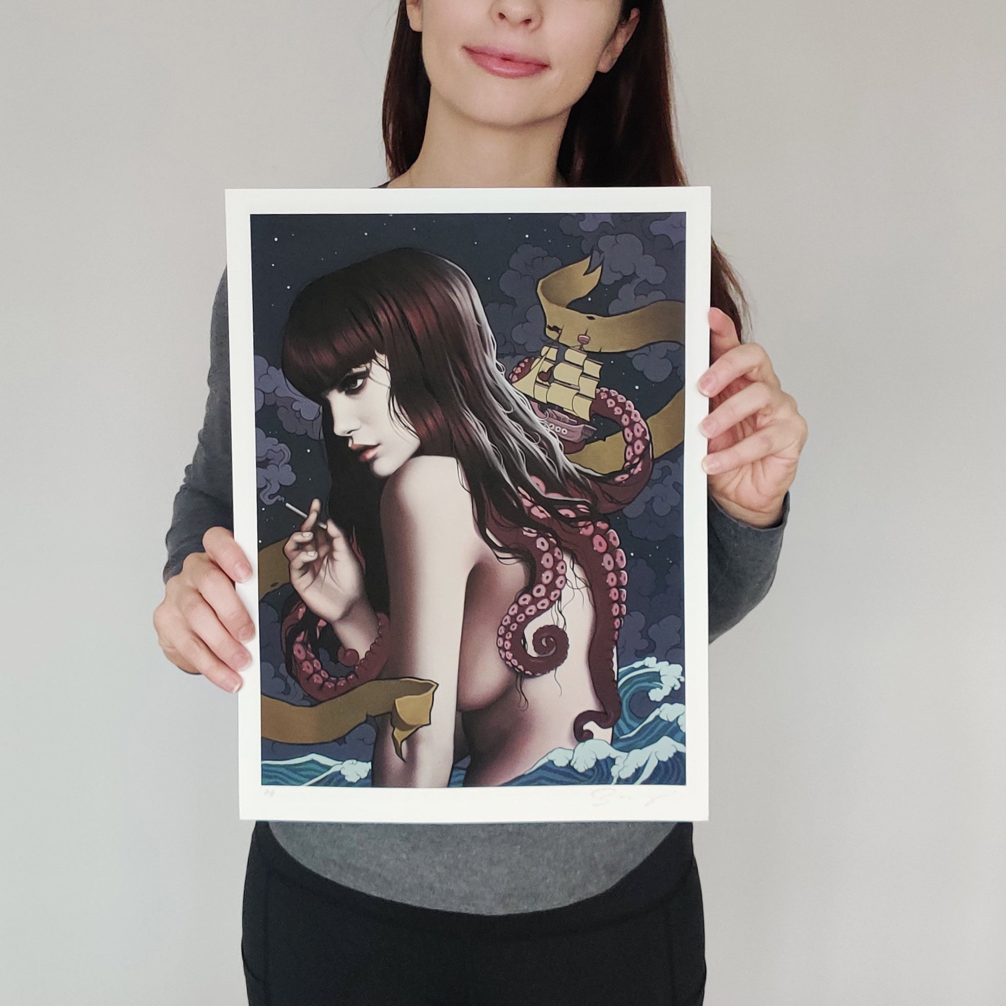 A person with long brown hair, in a gray top and black pants, holds Leviathan, an exquisite archival pigment print by Sarah Joncas. It illustrates a woman with long hair and an exposed back, entwined with tentacles against a night sky backdrop where a ship braves the sea.