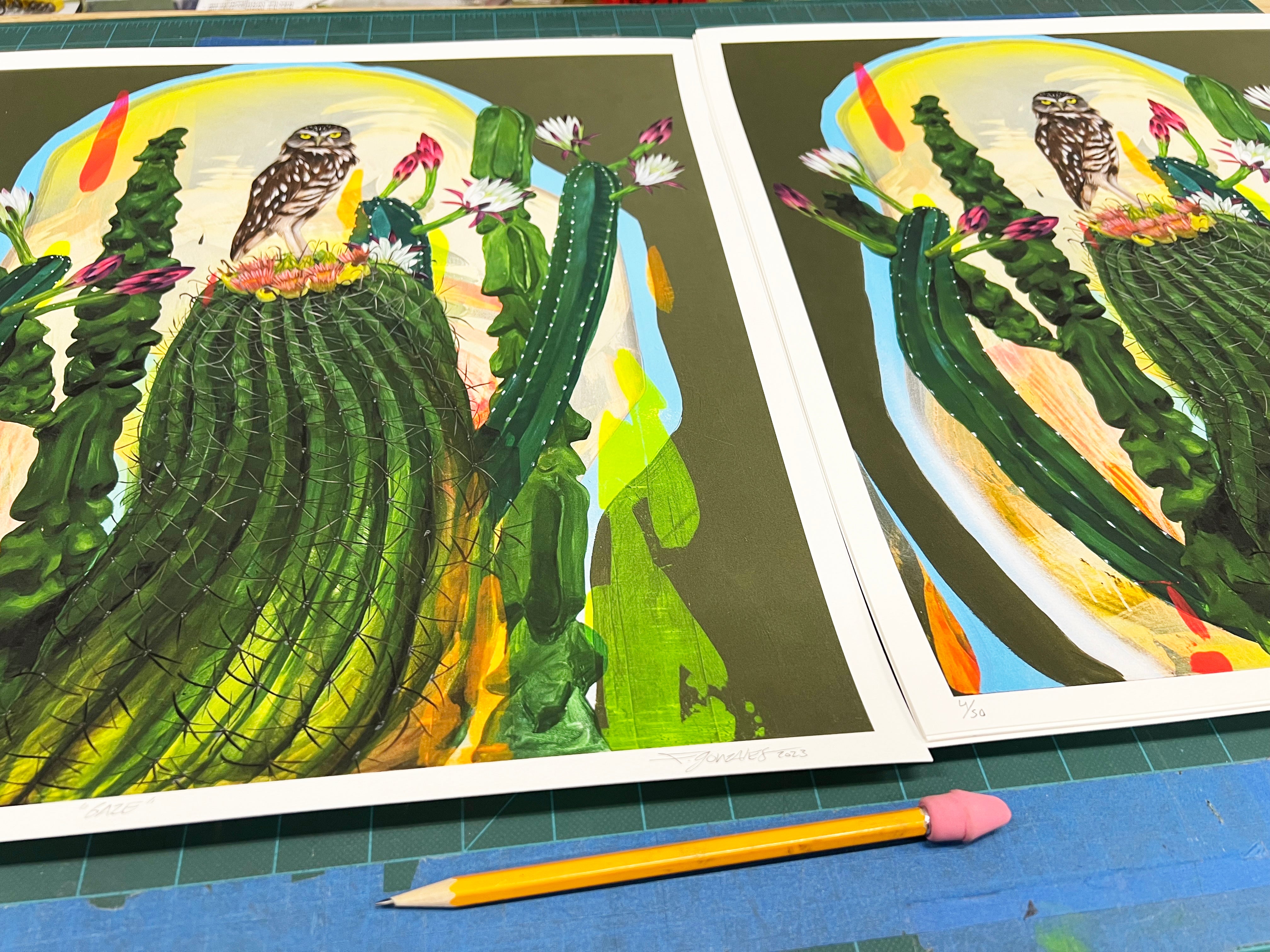 Two vibrant archival pigment prints titled Gaze by Frank Gonzales, signed and numbered in an edition of 50, depict an owl on a cactus among flowers. They are displayed on a green cutting mat next to a yellow pencil with a pink eraser.