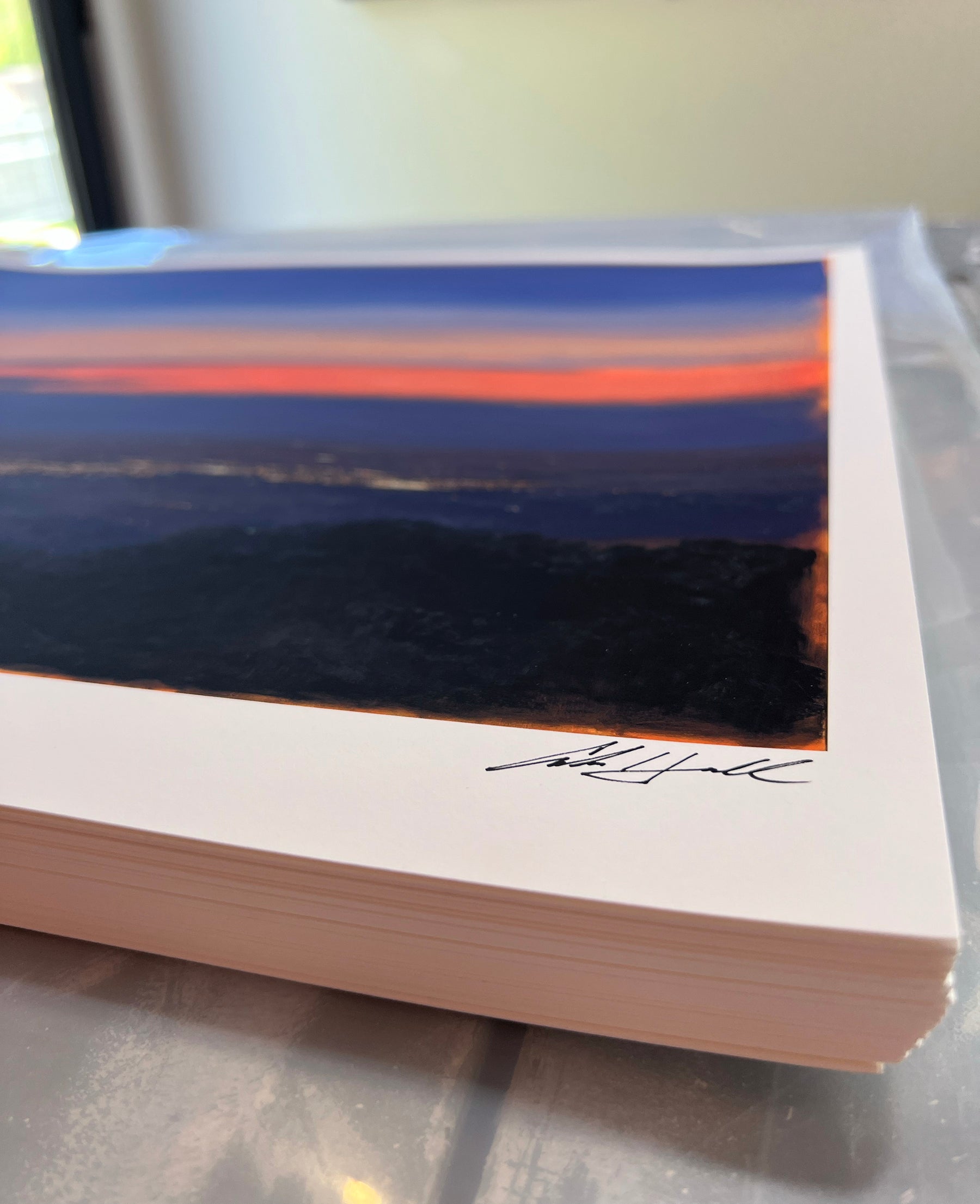 A stack of prints, featuring Nocturnal Valley by Adam Hall shows a landscape at sunset with orange and blue hues. The top print, an Archival Pigment Print, bears a signature on its edge. They rest on a plastic surface by a window in natural light.