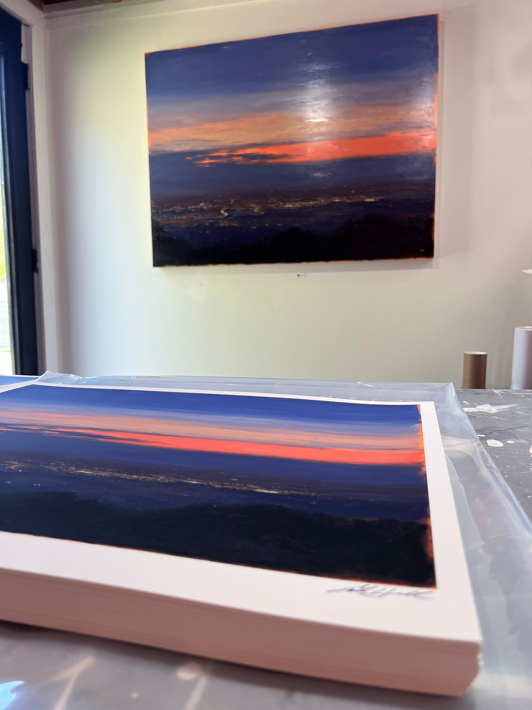 A photo captures two Adam Hall landscape artworks: Nocturnal Valley, with vibrant orange and blue hues. One artwork, an Archival Pigment Print, is mounted on the wall while the other is placed on a table in the foreground.