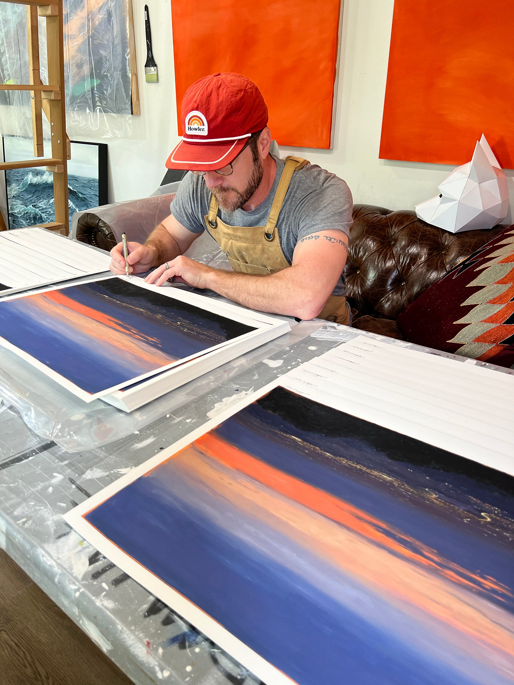 A person in a red cap and apron sits at a table, signing archival pigment prints of Adam Halls Nocturnal Valley, showcasing a vibrant sunset over a cityscape. The artistic space is decorated with colorful paintings and a geometric white wolf sculpture.