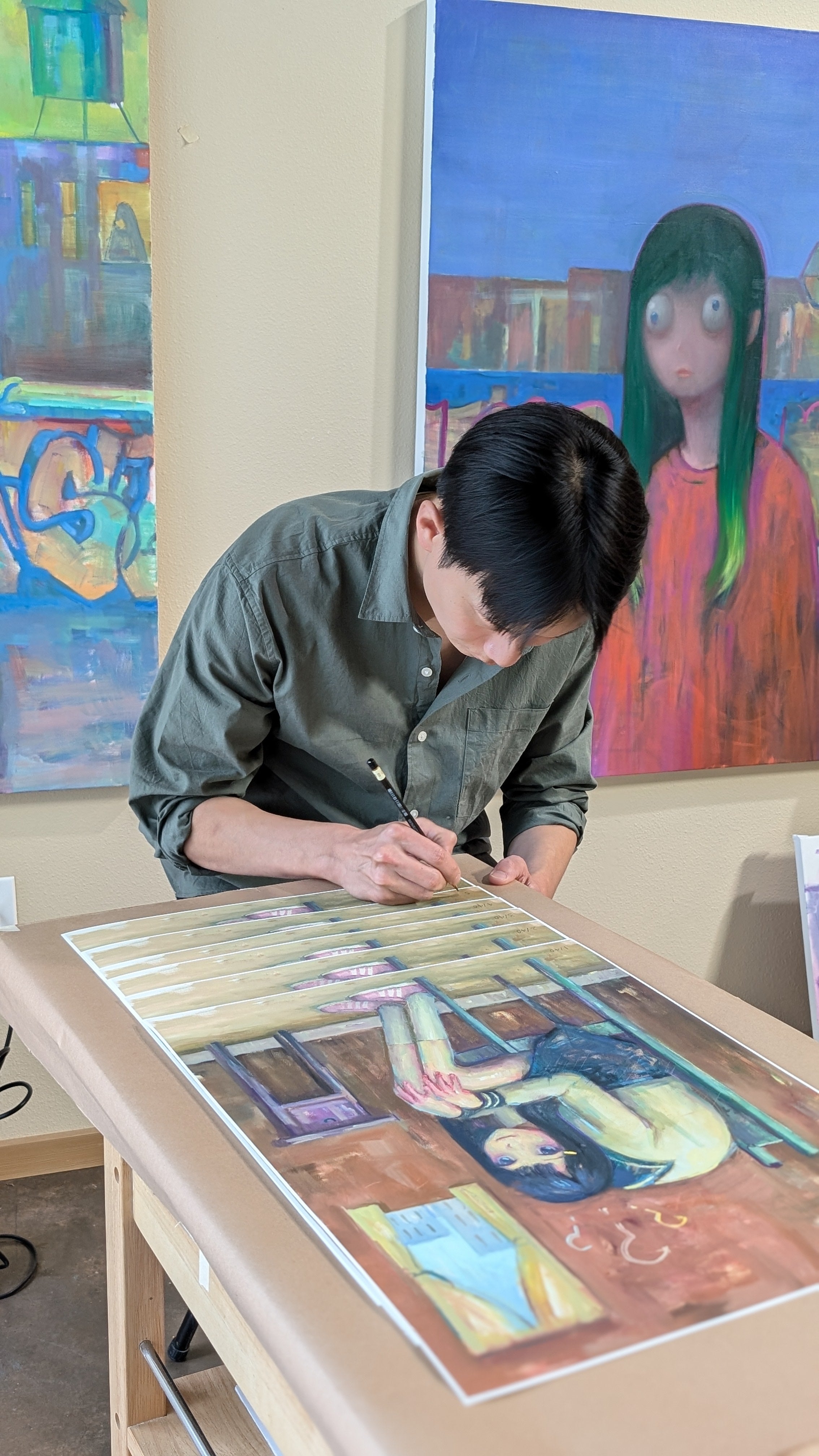 A person with short black hair bends over a table, creating a colorful painting of a contemplative figure. Surrounding them are vibrant artworks, including ??? by Leegan Koo, a signed and numbered limited edition archival pigment print.
