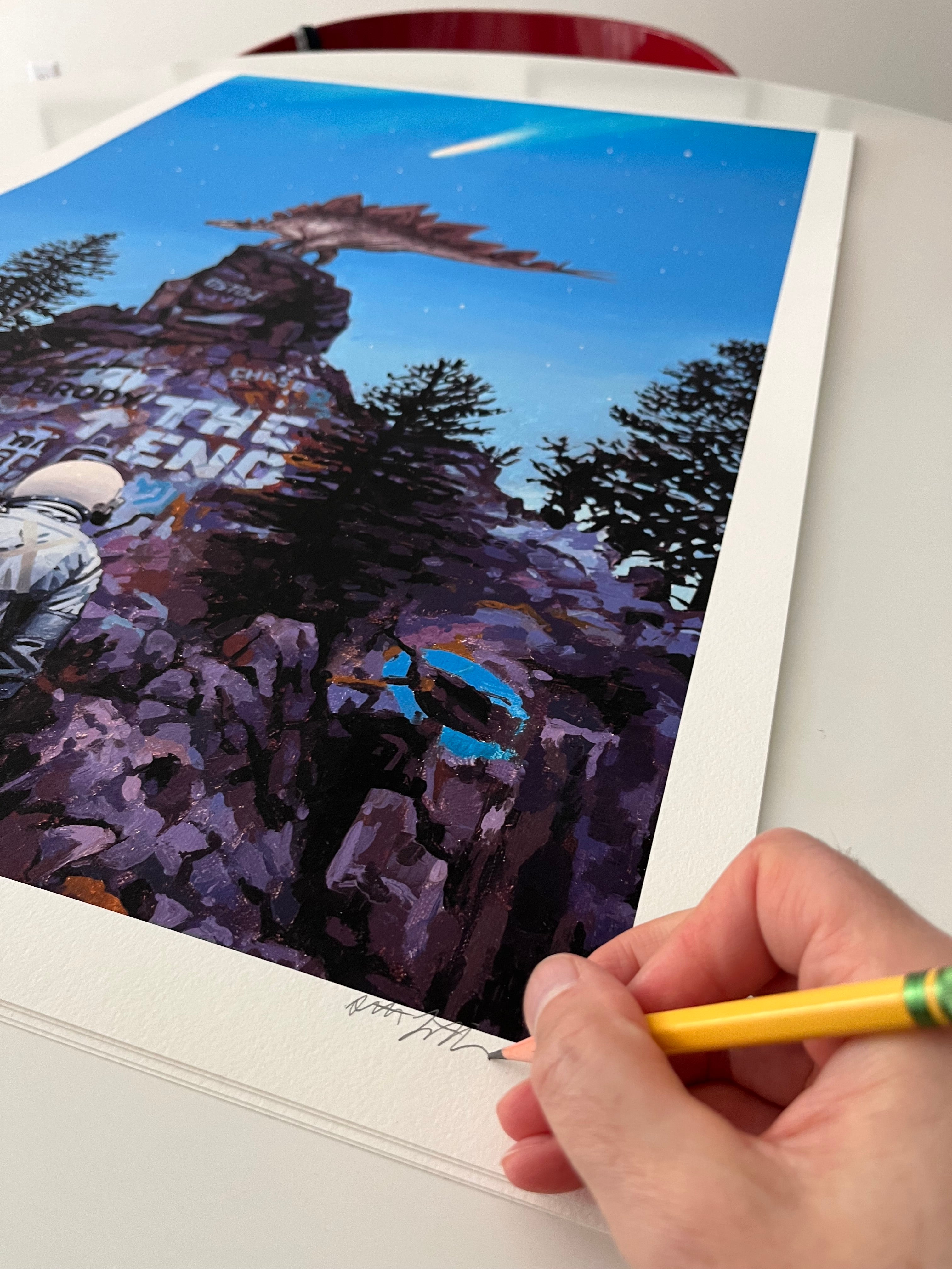 A hand holding a pencil signs "Stegosaurus 2," an archival pigment print by Scott Listfield, showing an astronaut, rocky cliffs, trees, and a large statue head beneath a starry blue night sky on a white table.