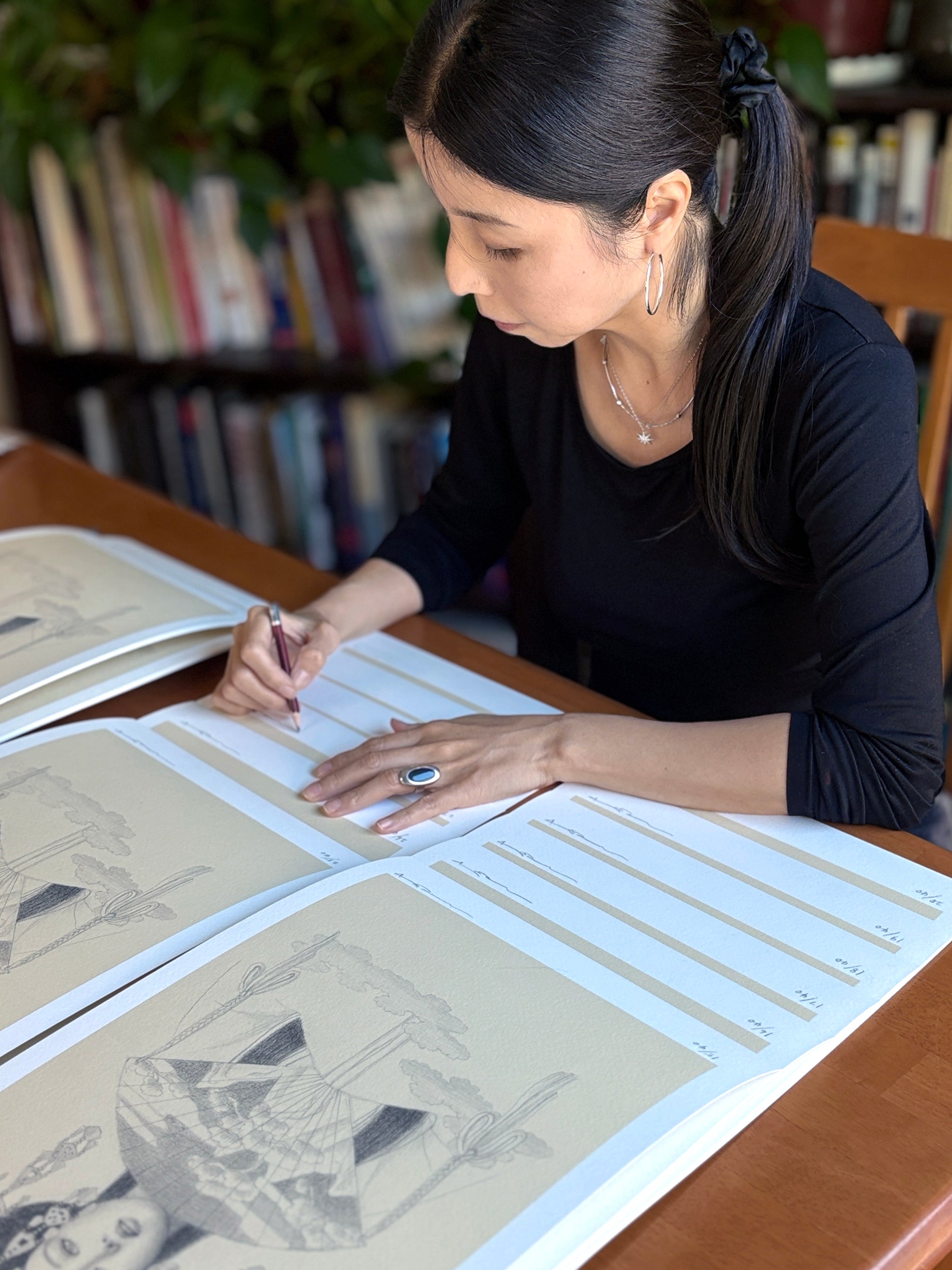 A woman with long black hair in a ponytail signs limited edition Hemera archival pigment prints by Chie Yoshii at a table covered with more prints. Bookshelves and green plants are visible in the background.