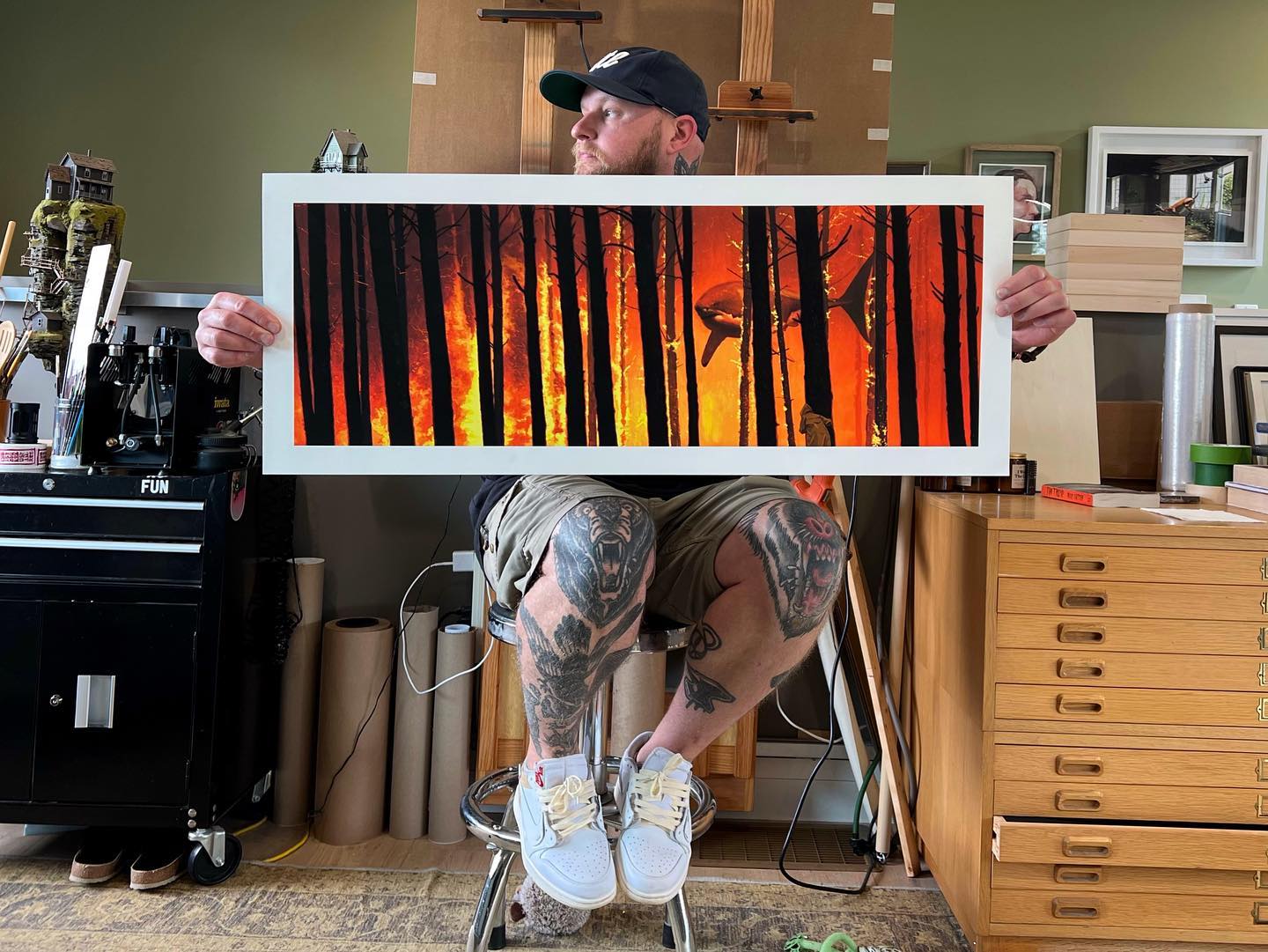 A tattooed man sits on a stool in a studio filled with art supplies, holding "No End In Sight," a wide Chris Austin archival pigment print depicting black tree silhouettes and orange flames of a forest fire.
