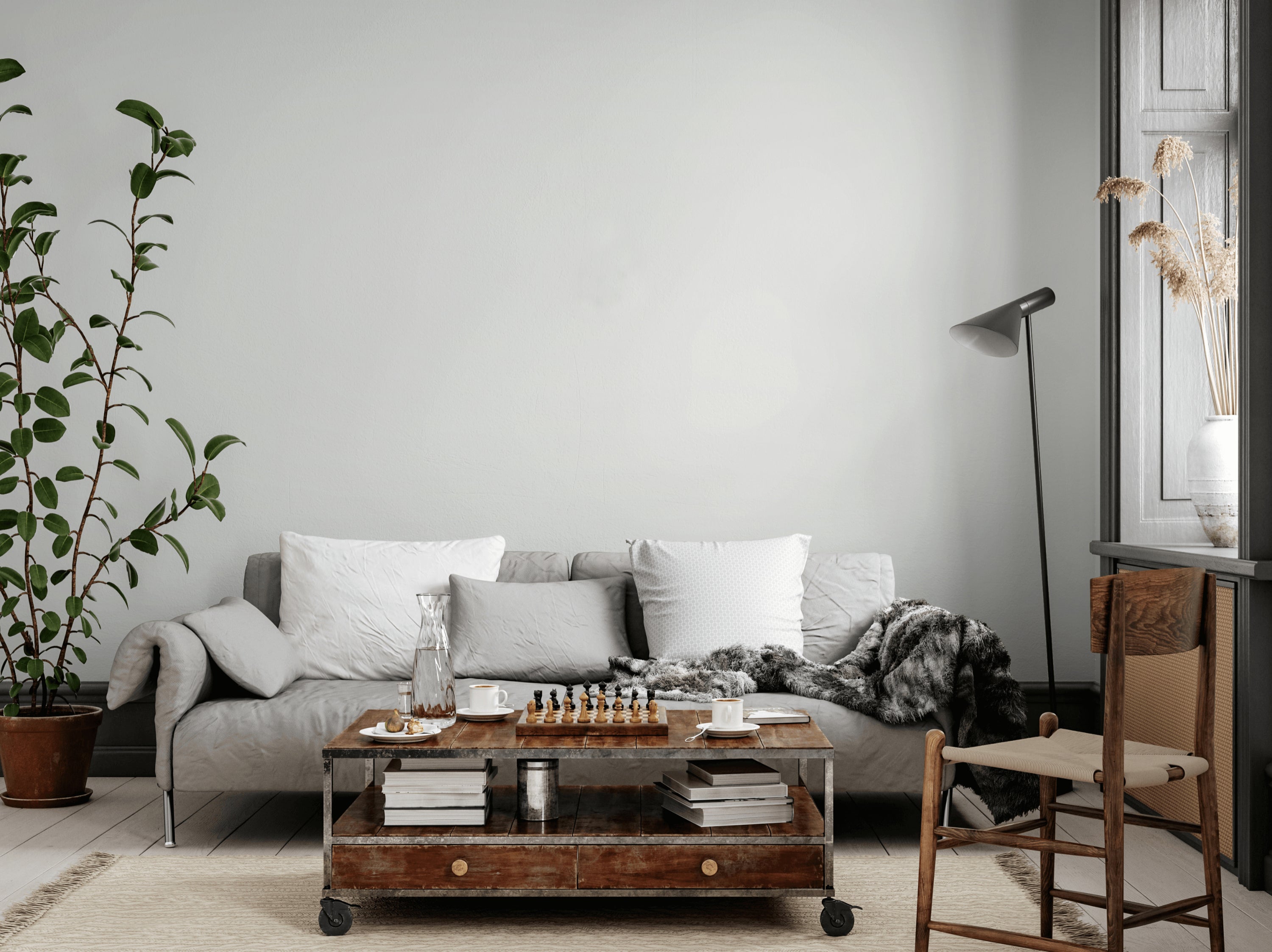 A cozy living room with a light gray sofa adorned with white pillows and a plush throw. A rustic wooden coffee table displays a chess set and glasses, under a floor lamp. A tall plant and wooden chair add to the decor, with natural light from the window.