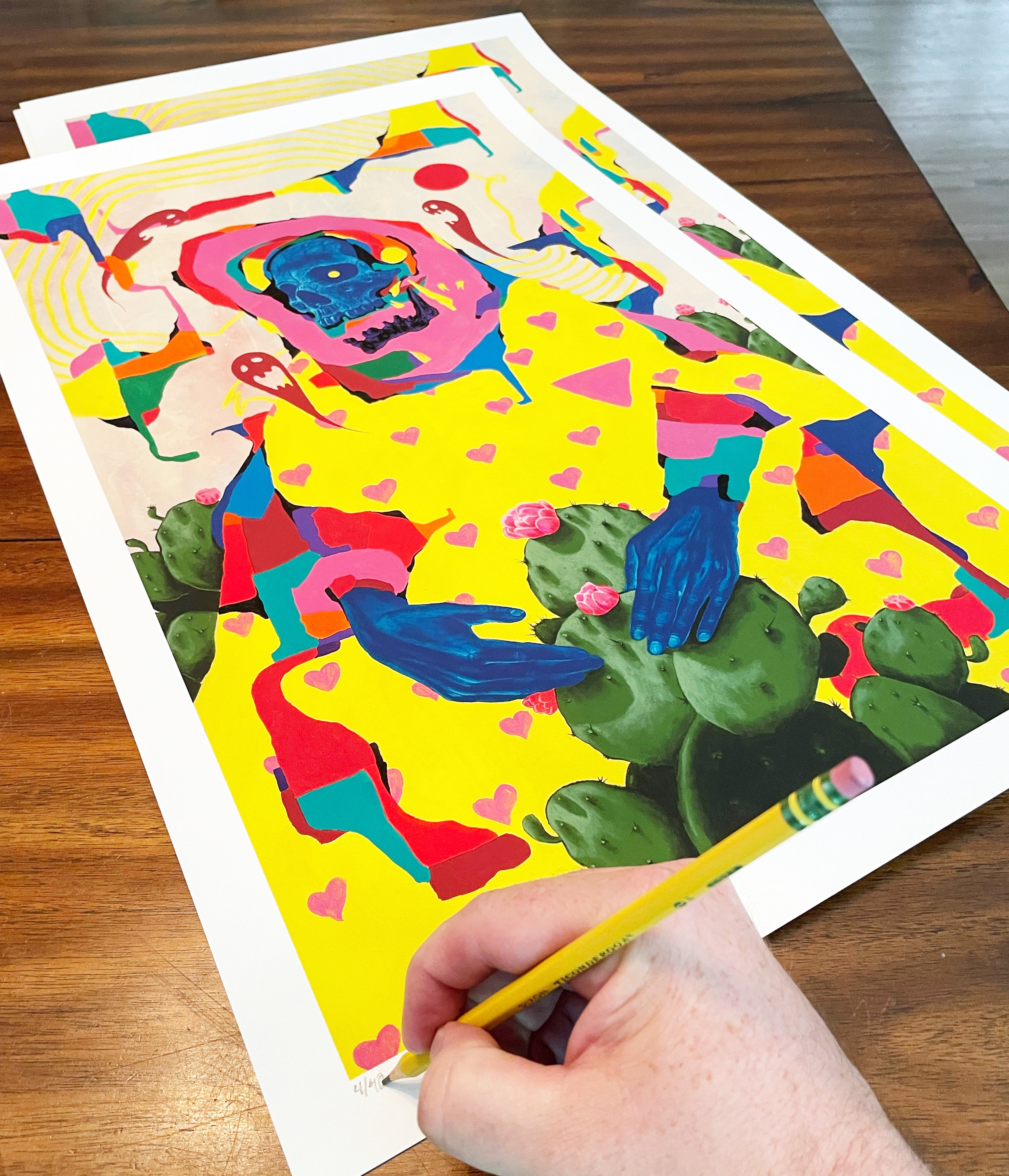 A hand signs and numbers the “What Doesn’t Kill Us” archival pigment print by Anthony Hurd, showing a blue-faced figure with blue hands, hearts, and cacti on a bright yellow background atop a wooden table.