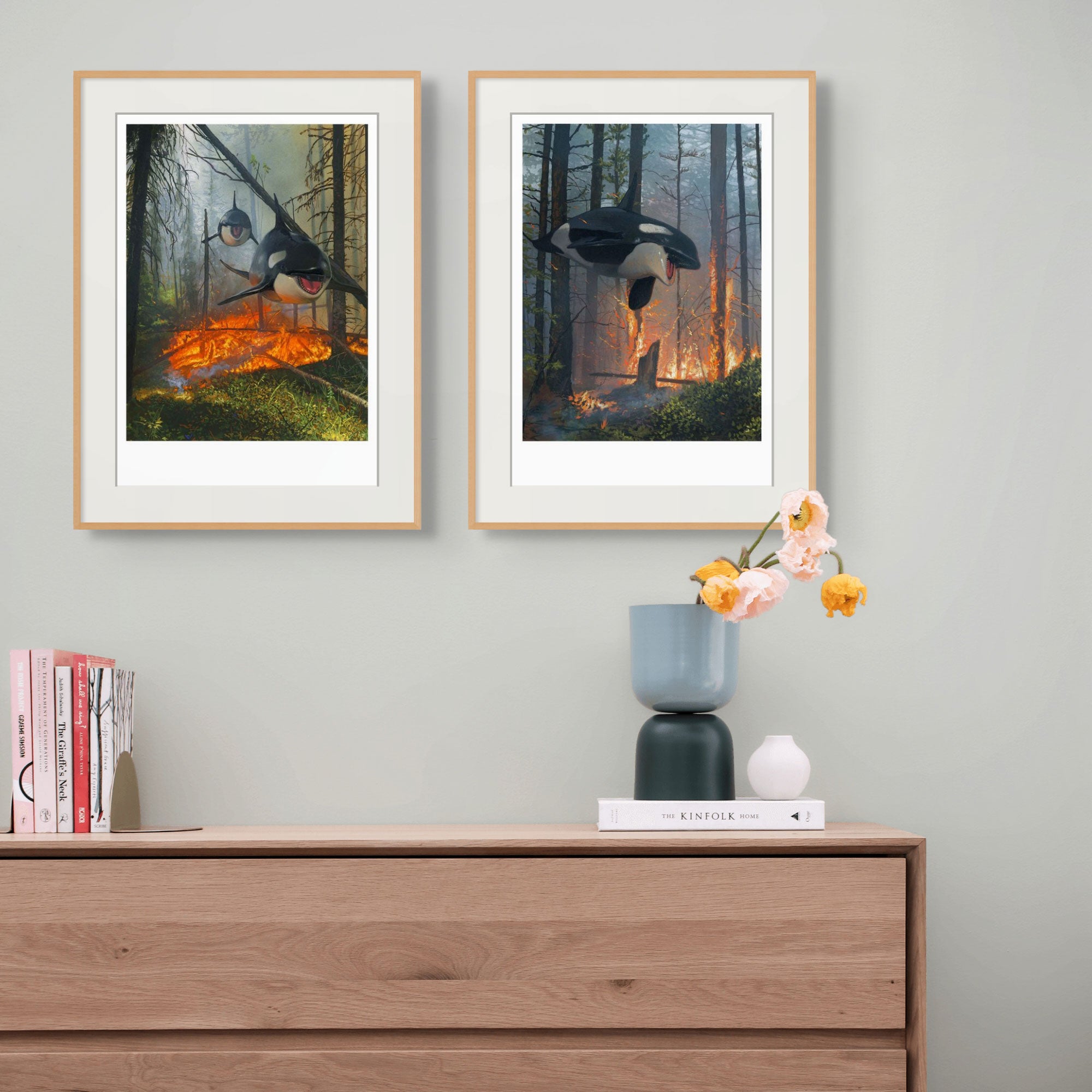 A wooden dresser with a black and white vase holding pink flowers is in front of a light green wall. Two framed artworks of orcas in a forest with a fire hang above. A few books are stacked on the left side of the dresser.