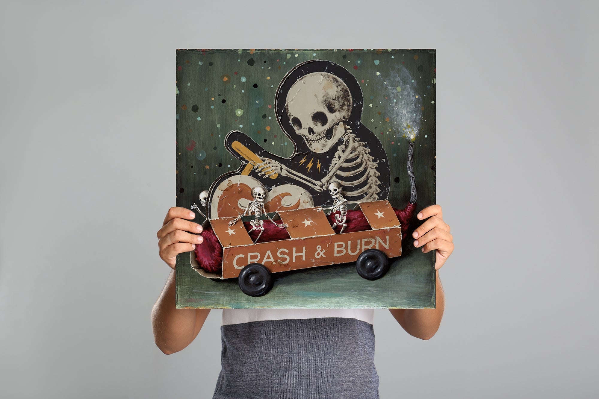 Someone holds up the "Crash & Burn" painting by Jason Limon, featuring a skeleton in a toy car, obscuring their face behind the artwork against a plain gray background.