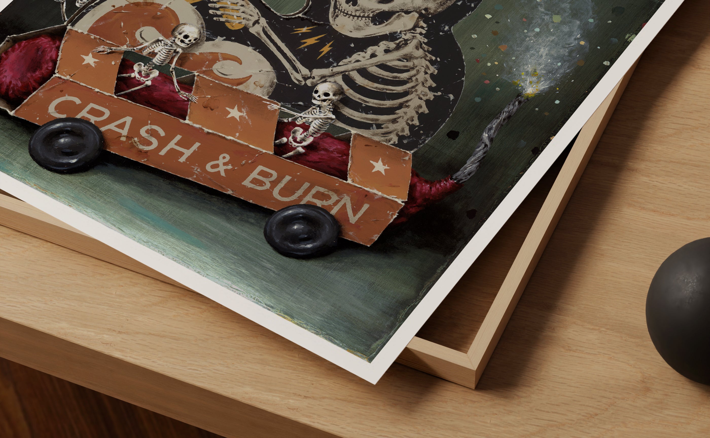 A close-up of Jason Limon's "Crash & Burn" Archival Pigment Print shows skeletons in a cardboard car labeled Crash & Burn with stars, set against a vivid cosmic backdrop on wood.