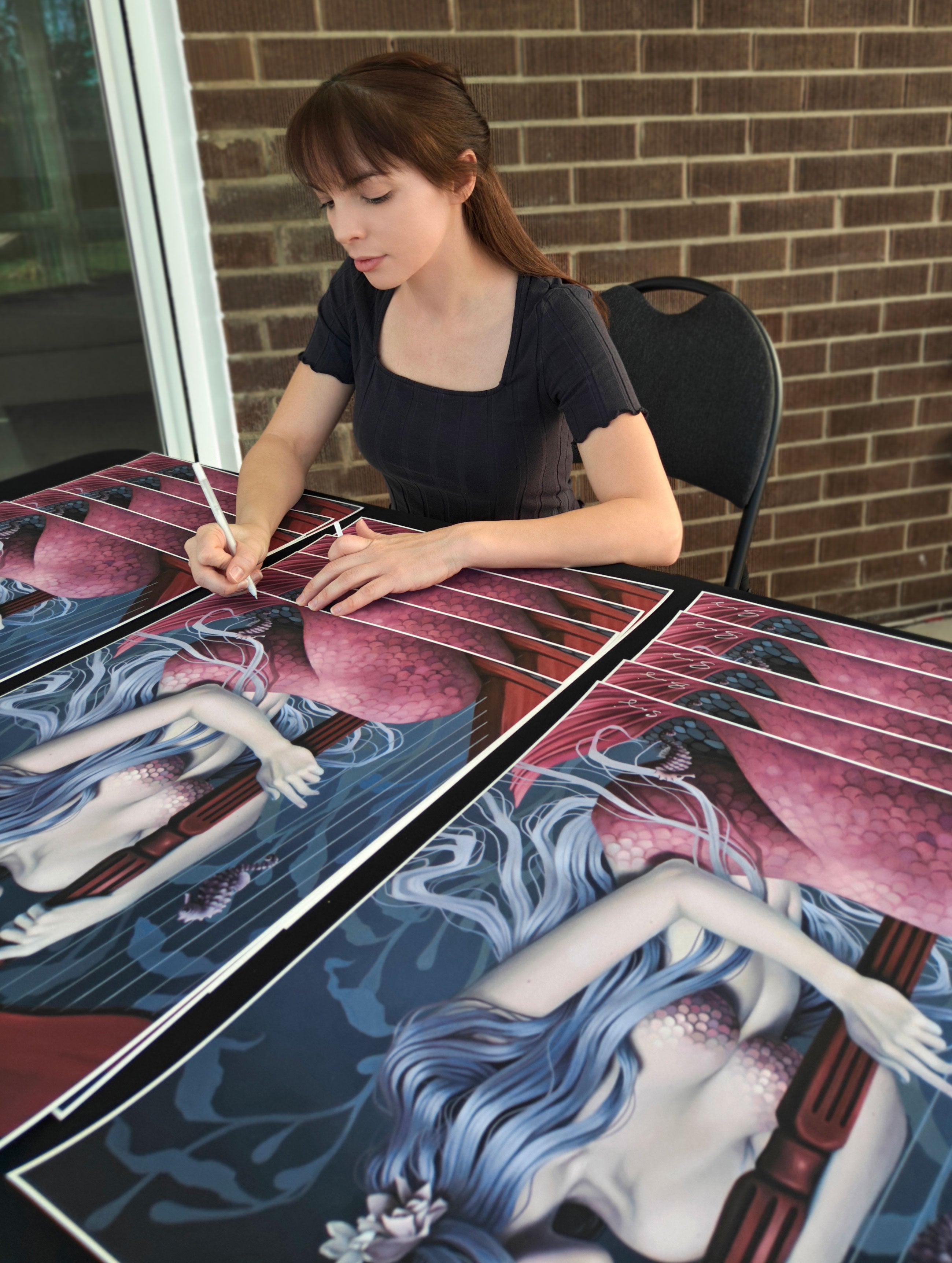 Clad in a black dress, a person intently signs archival pigment prints of Sea Sonata by Sarah Joncas, depicting a mermaid with blue hair and a pink tail. The artistic scene is set against a backdrop of a brick wall and window.