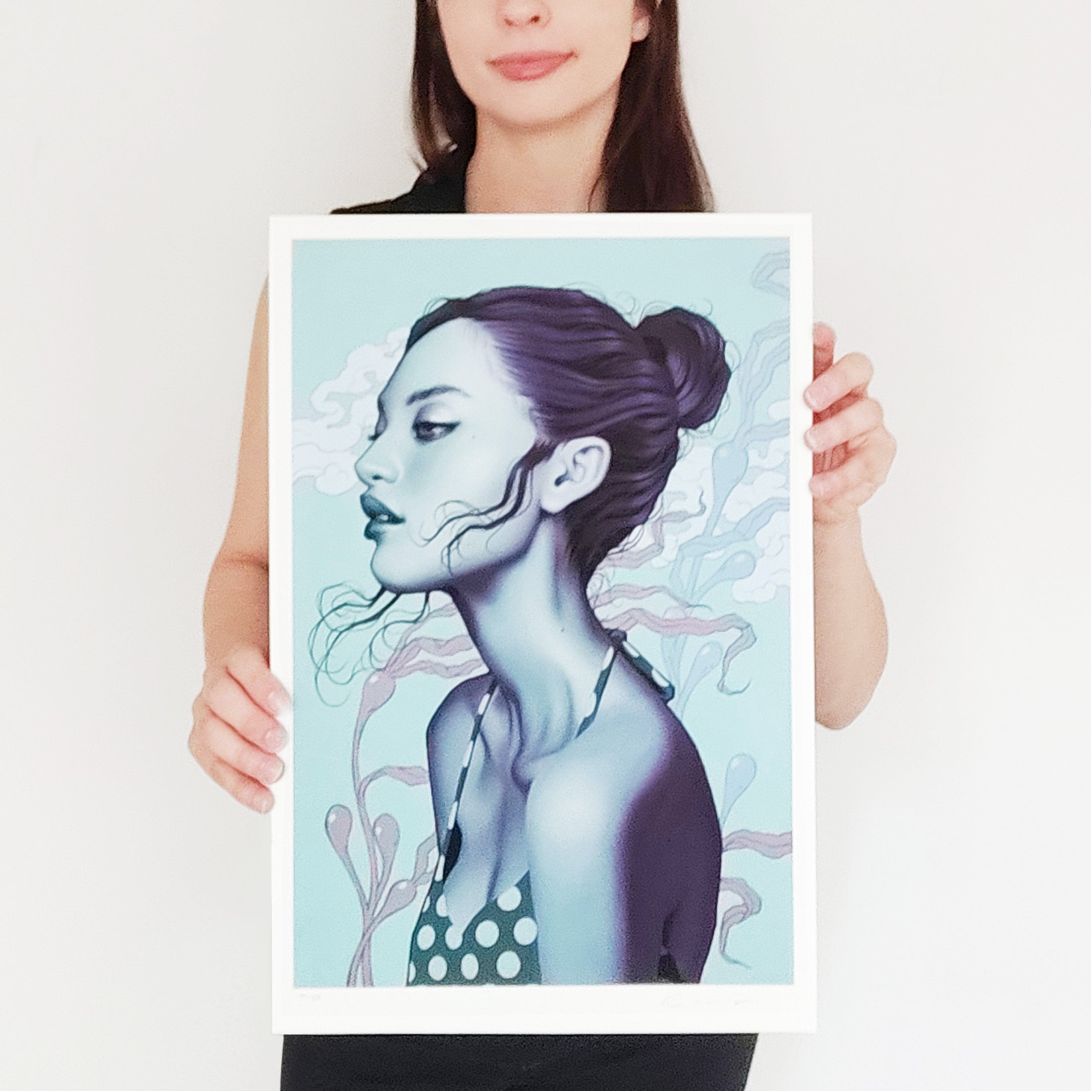 A person holds Sea Breeze, an archival pigment print by Sarah Joncas. It features a stylized portrait of a woman with dark hair in a bun, wearing a polka-dot top against a light blue background with abstract patterns.