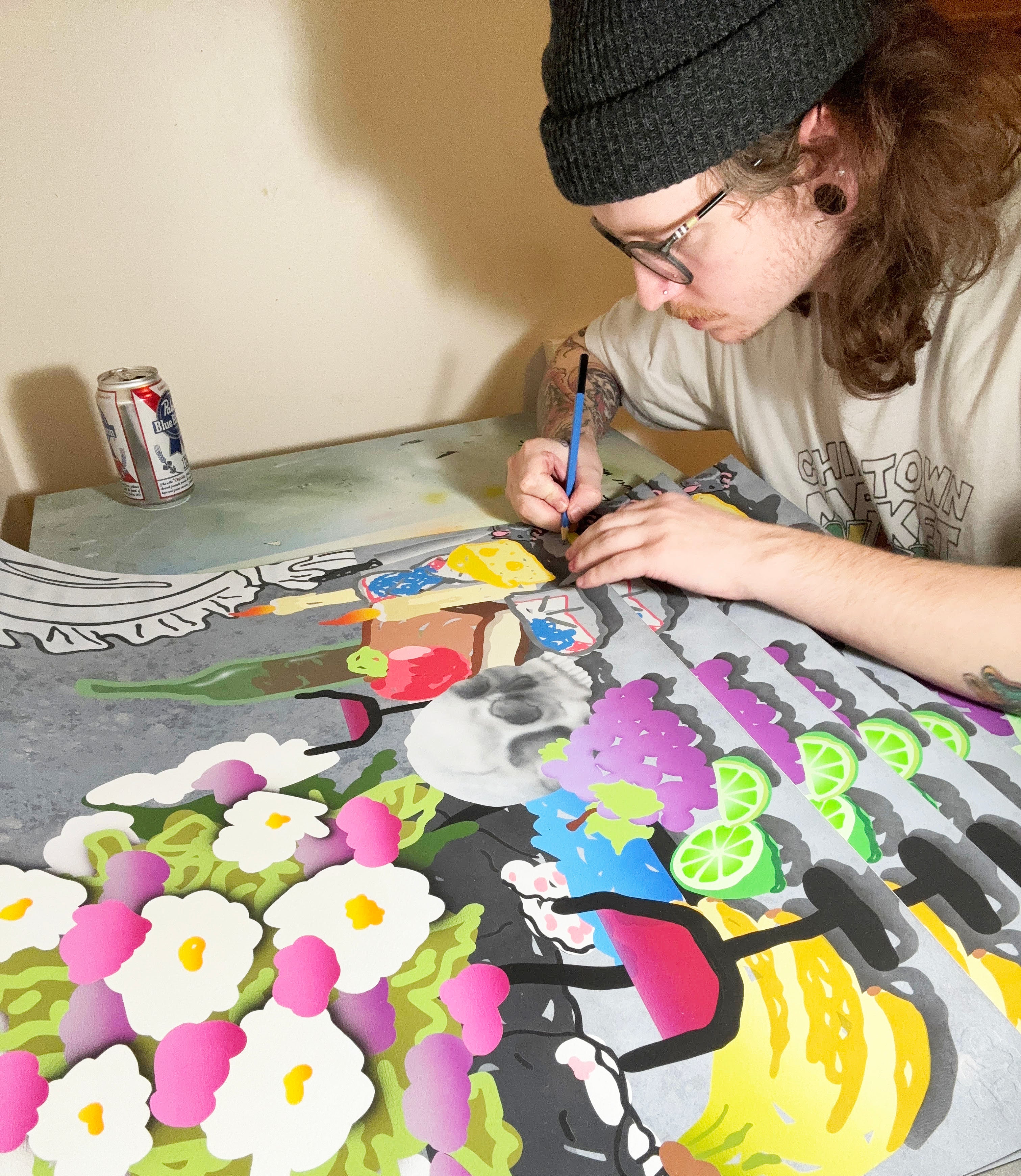 Wearing glasses and a beanie, an artist draws a vivid still life of flowers and fruit beside a can of soda, inspiring "Still Life Study"—an archival pigment print by Jordan Ferguson.