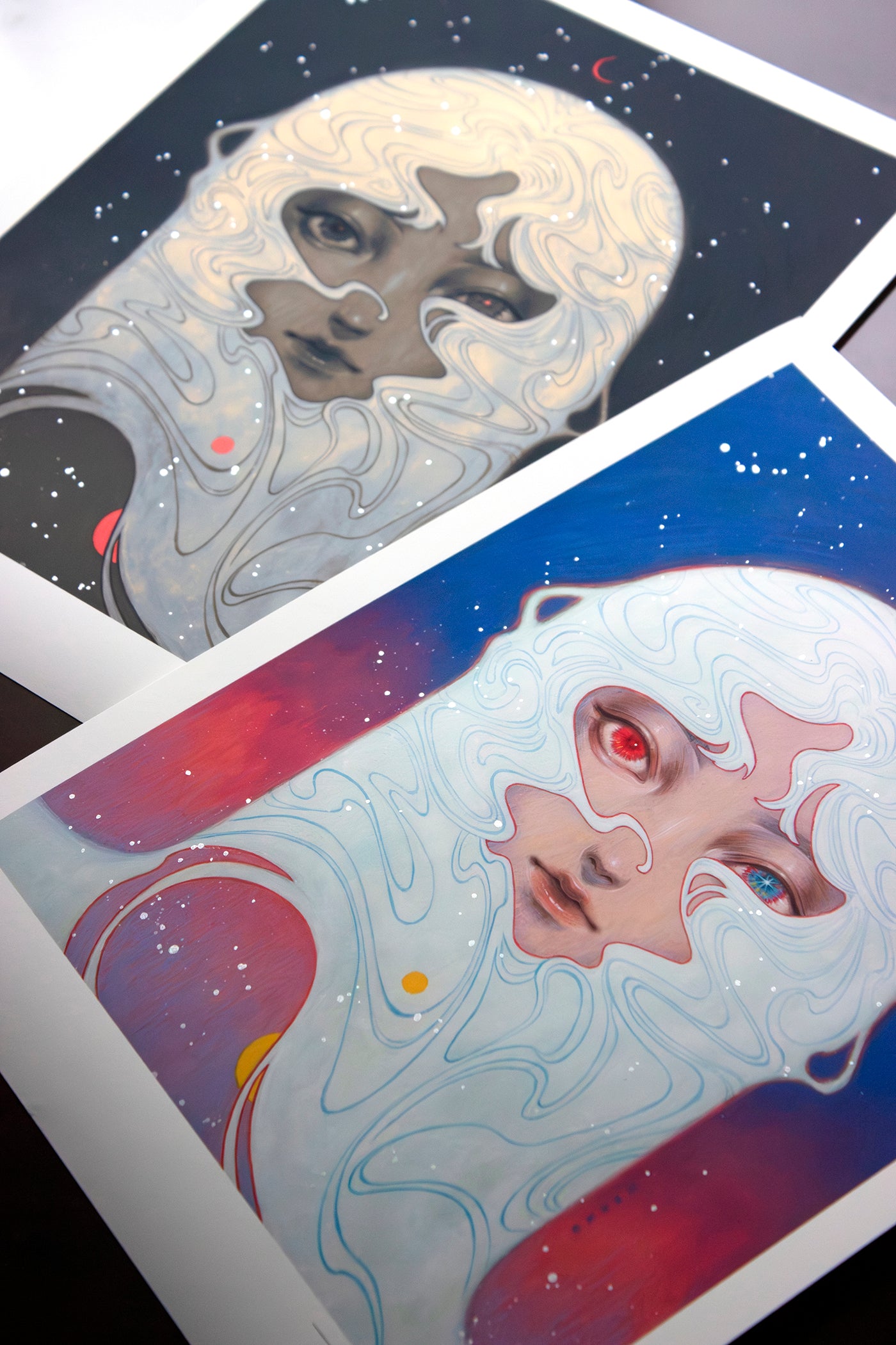Two art prints by Shoko Ishida feature abstract portraits with swirling white patterns: the top, a Hand Embellished Edition with a dark background for a serene feel; the bottom, Melody of Dawn, an Archival Pigment Print on blue, depicting one red and one blue eye.