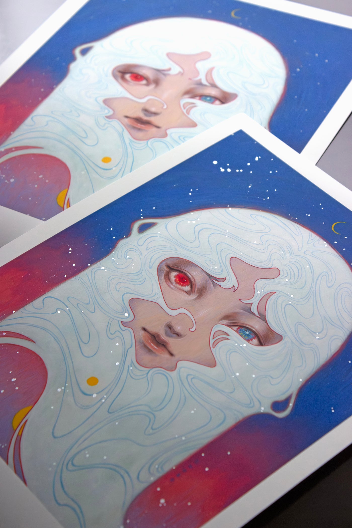 Two Melody of Dawn art prints by Shoko Ishida feature a stylized face with mask-like elements, mismatched red and blue eyes, and create an otherworldly scene with a blue-pink gradient, star dots, and a crescent moon in archival pigment print.