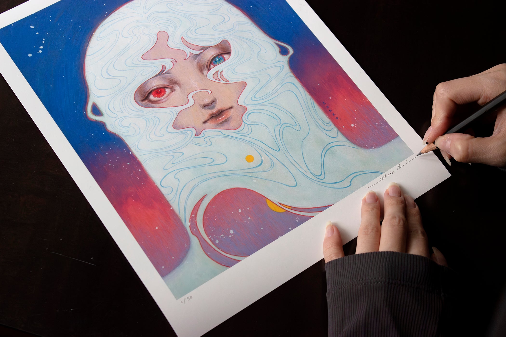 An individual delicately signs a Melody of Dawn art print by Shoko Ishida, part of the Hand Embellished Edition, showcasing a surreal face with red eyes, framed in white lines on a blue and pink gradient.