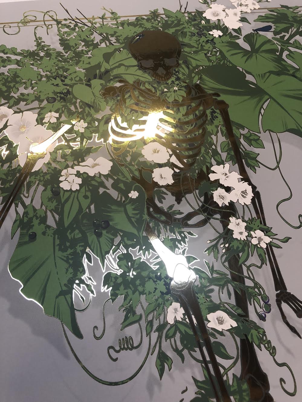 The Fertile Ground screenprint by JAW Cooper features a skeleton entwined with lush green leaves and white flowers, intricately detailed with a reflective sheen against a neutral background, evoking the fertile ground of imagination.