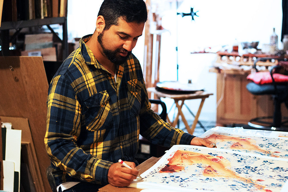 A man in a plaid shirt works intently in an art studio, marking floral-printed paper titled Thisbe, crafted by Sergio Lopez. Each piece is part of a 40-edition series as an archival pigment print, signed and numbered, with blurred shelves and chairs in the background.