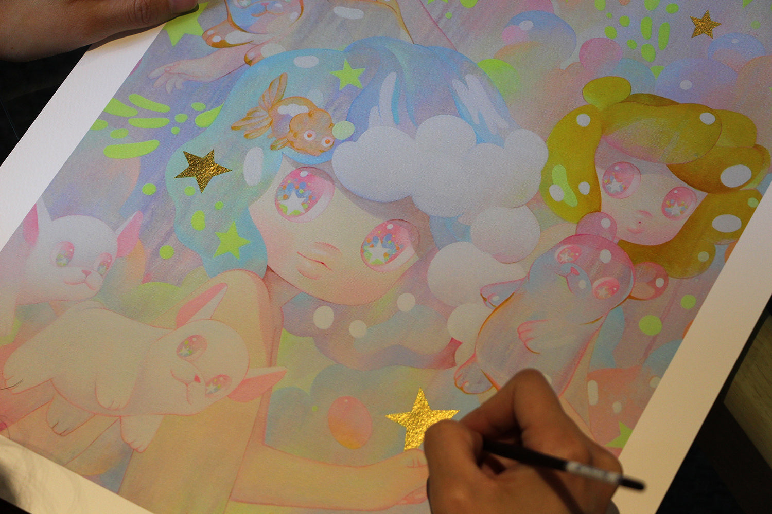 An individual is using a black pen to hand embellish Lullaby by So Youn Lee, enhancing whimsical characters like a blue-haired girl, cute animals, and small fish on this pastel-hued archival pigment print adorned with golden stars.