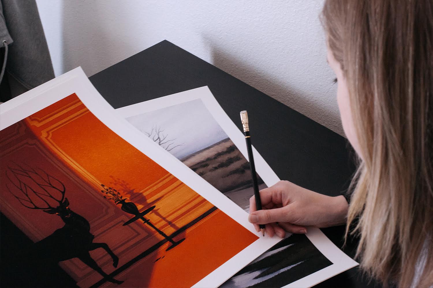 A light brown-haired person with a pencil inspects two art prints on a black table: an Archival Pigment Print by Jenna Barton showcasing an orange scene with a deer silhouette, and Darker Rooms, a muted landscape ideal for dimly lit spaces.