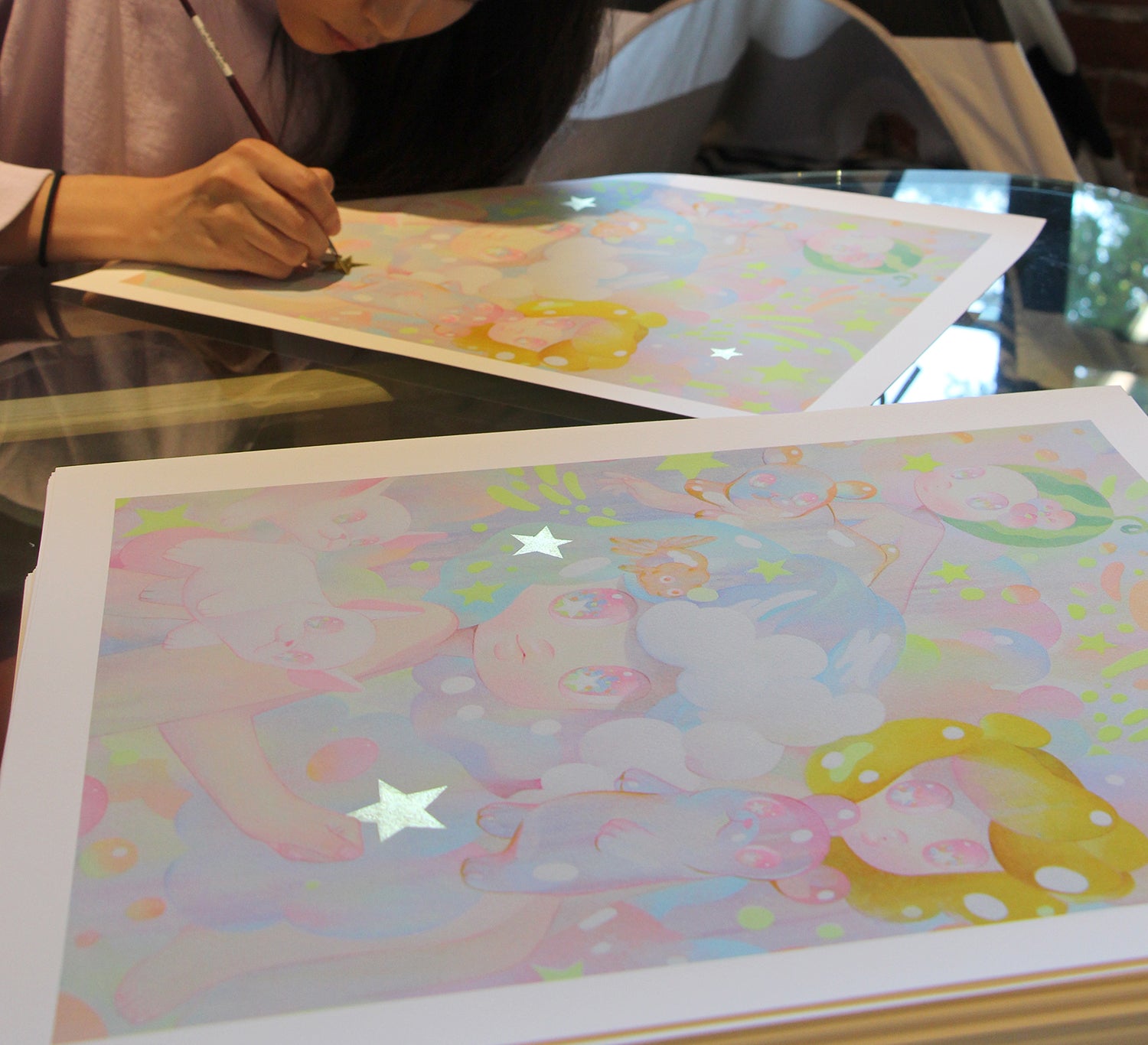 A person carefully signs a colorful print titled Lullaby by So Youn Lee, showcasing dreamy characters and shimmering stars. This archival pigment print lies on a glass table, highlighting intricate details, soft pastel hues, and hand-embellished accents.