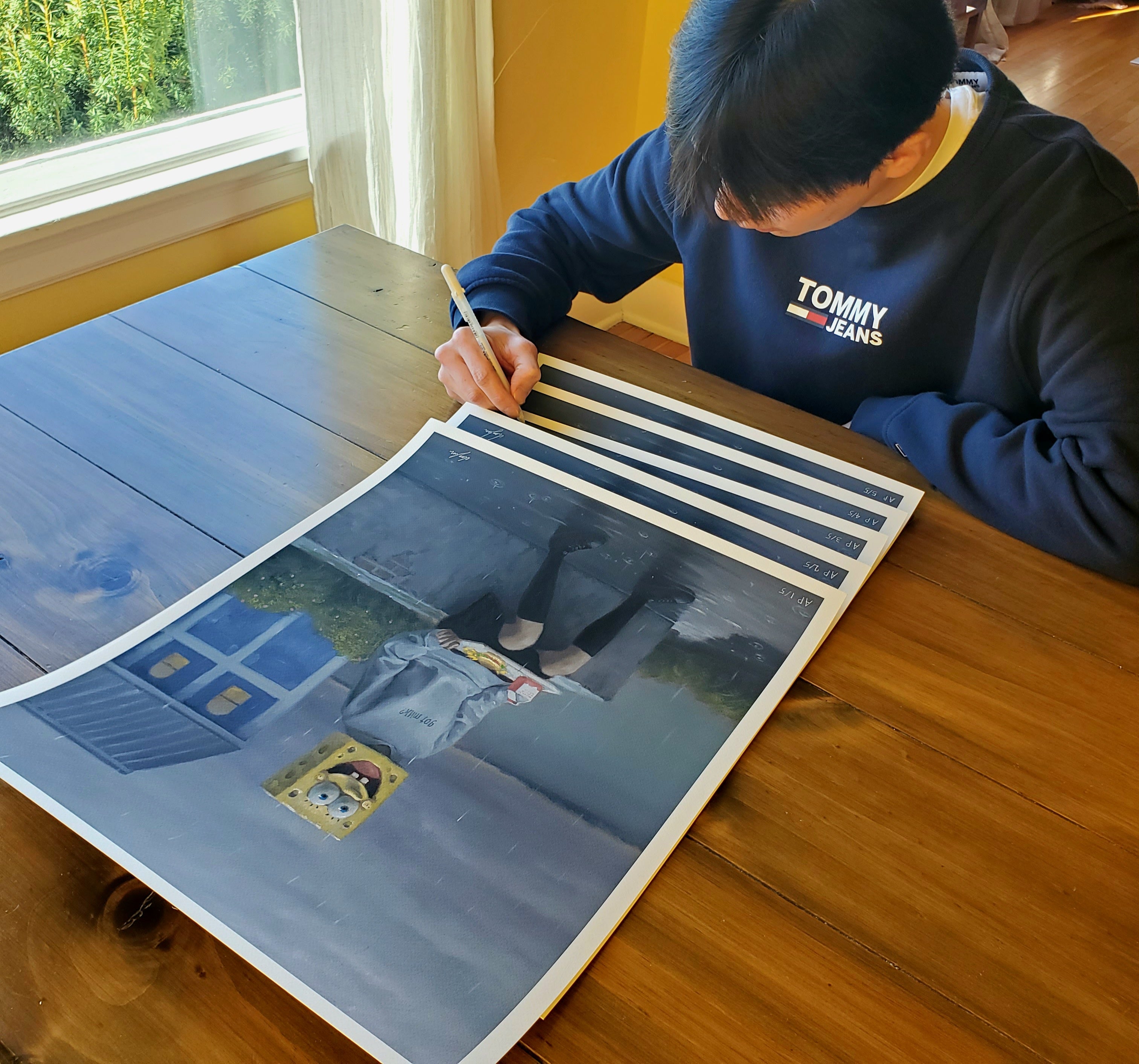 At a wooden table, someone in a navy sweatshirt signs and numbers large, limited edition prints of Lunch Period, an artwork by Leegan Koo featuring a character with a box-shaped helmet. Natural light from the left window highlights each archival pigment print.