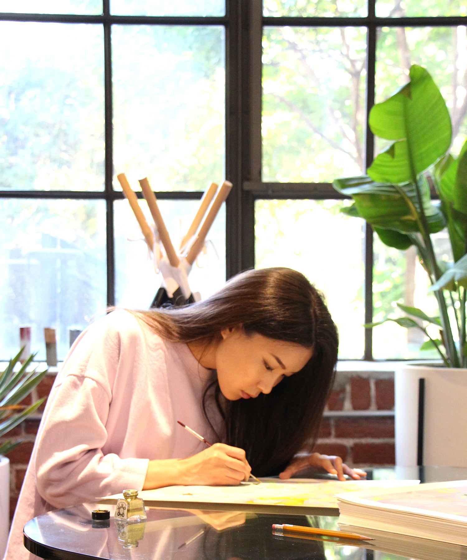 A woman with long dark hair sits at a glass table, focused on hand embellishing an art piece titled Lullaby by So Youn Lee. She wears a light pink sweater in a room adorned with brick walls, large windows, and lush plants filtering abundant natural light.