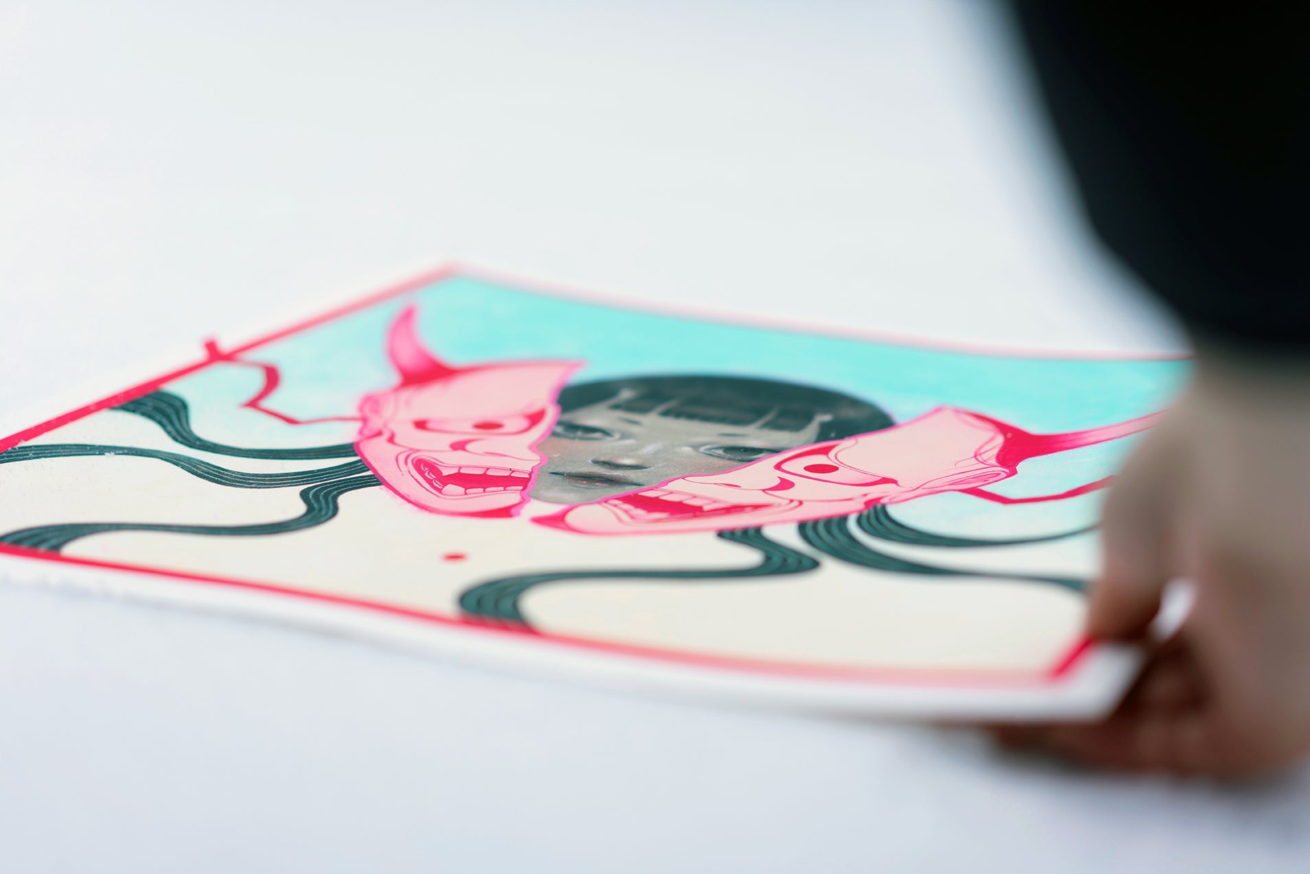 A person holds "HANNYA" by Shoko Ishida—an archival pigment print showing a woman's face with black hair, two pink HANNYA masks, and wavy black lines on a light blue background.