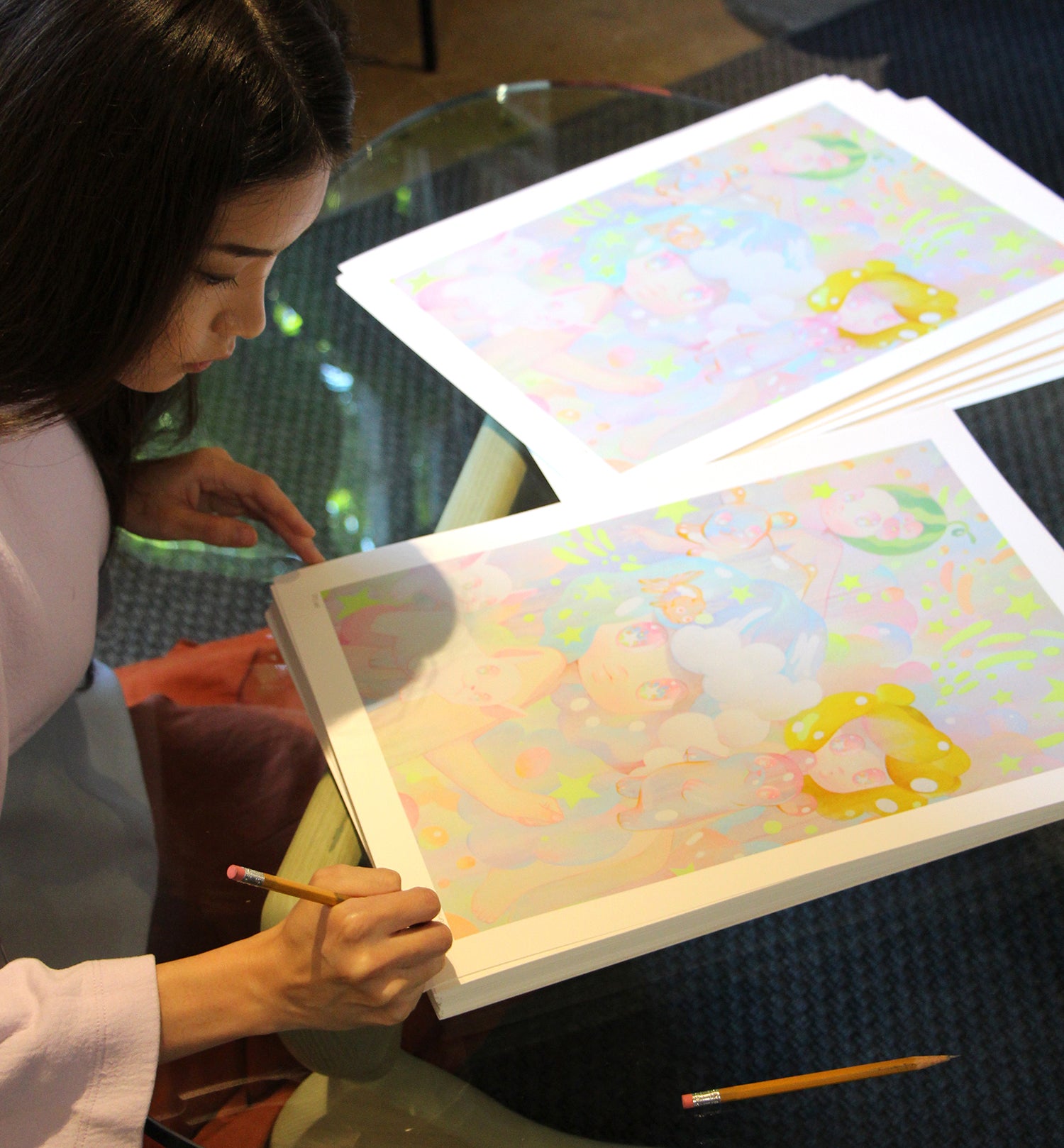 At a glass table, someone holds a pencil, examining So Youn Lees colorful Lullaby art prints. The artwork showcases pastel characters in vivid detail, and the reflective table surface highlights these hand-embellished masterpieces created with archival pigment prints.