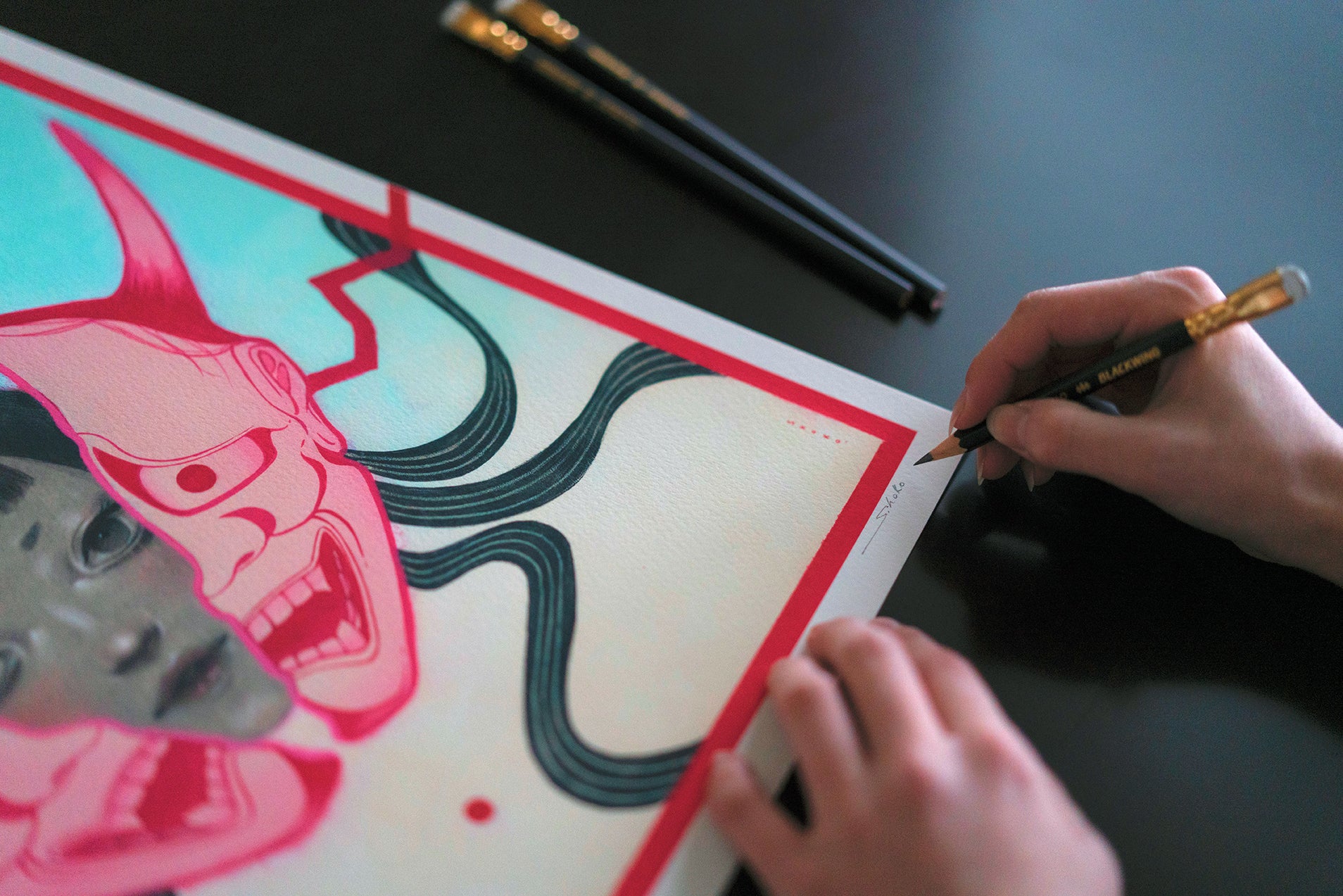 A person signs the corner of "HANNYA," a colorful archival pigment print by Shoko Ishida featuring expressive faces and flowing lines, using a pencil. Two pencils rest on a dark table in the background.