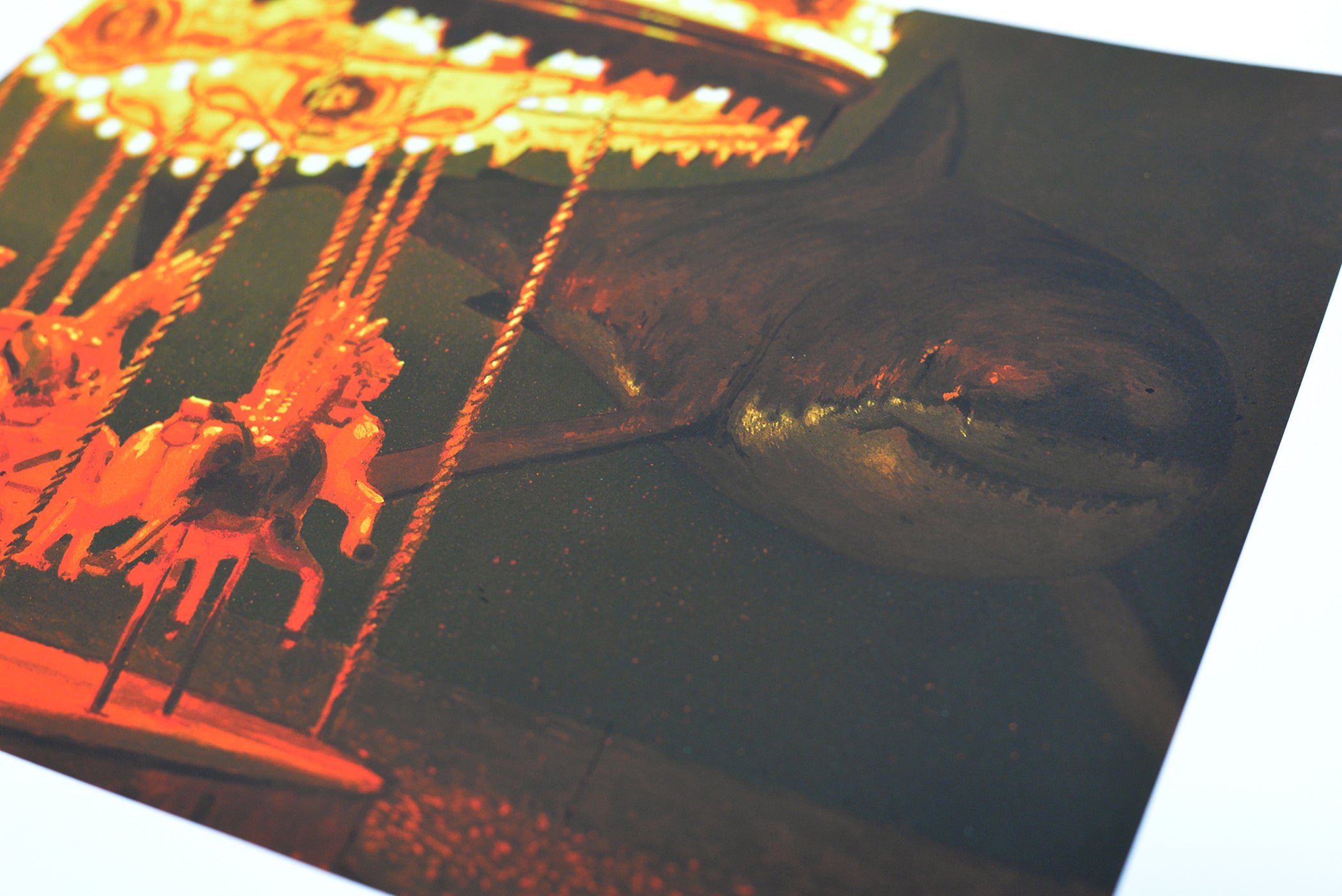 The archival pigment print Take Me To The Carnival by Chris Austin shows a dark underwater scene with a shark lurking near an eerie, glowing carnival carousel, where illuminated horses contrast with the nearby predator.