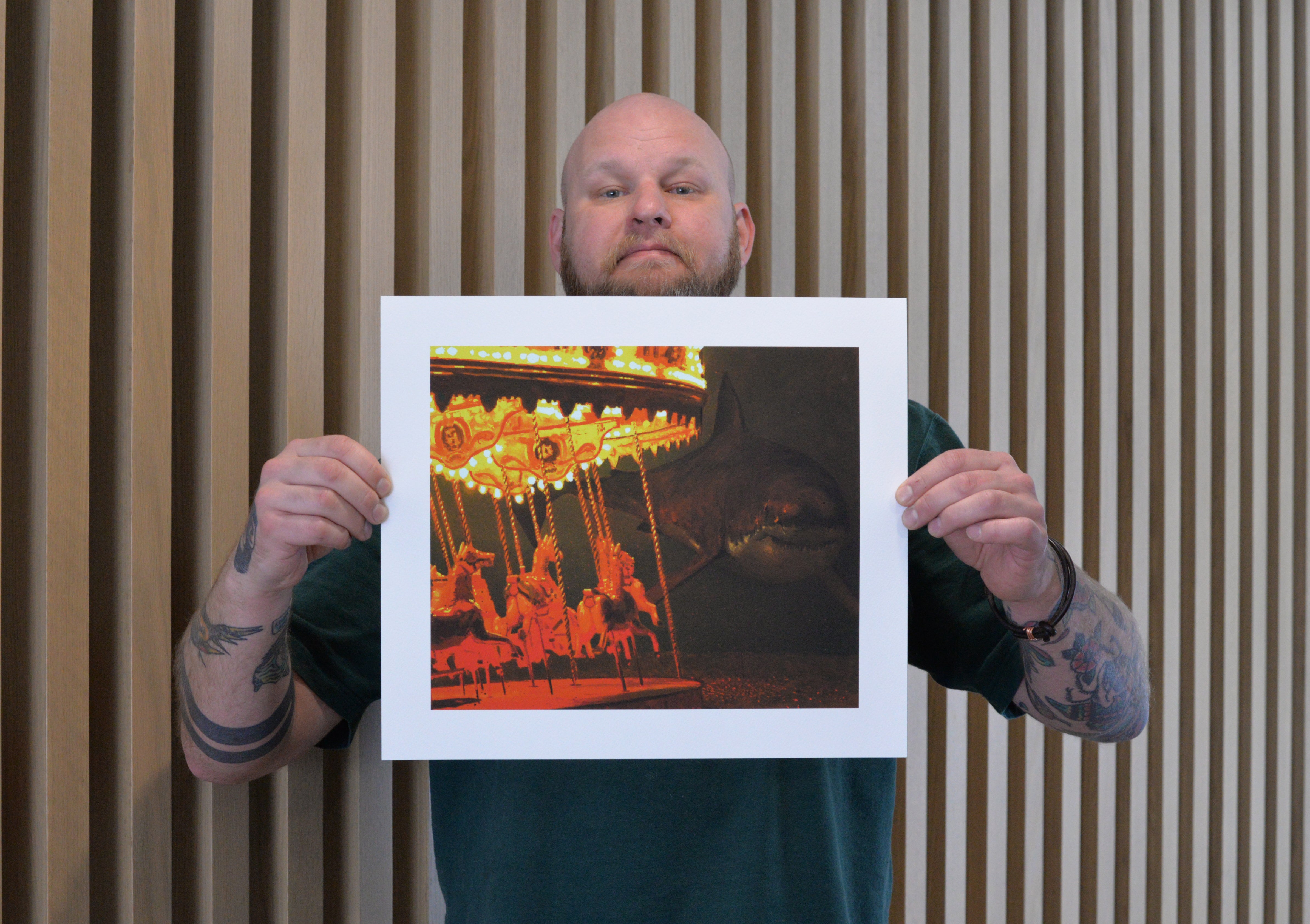 A tattooed person showcases Take Me To The Carnival by Chris Austin, an archival pigment print capturing a brightly lit carousel with red lights and horse figures against vertical wooden slats.