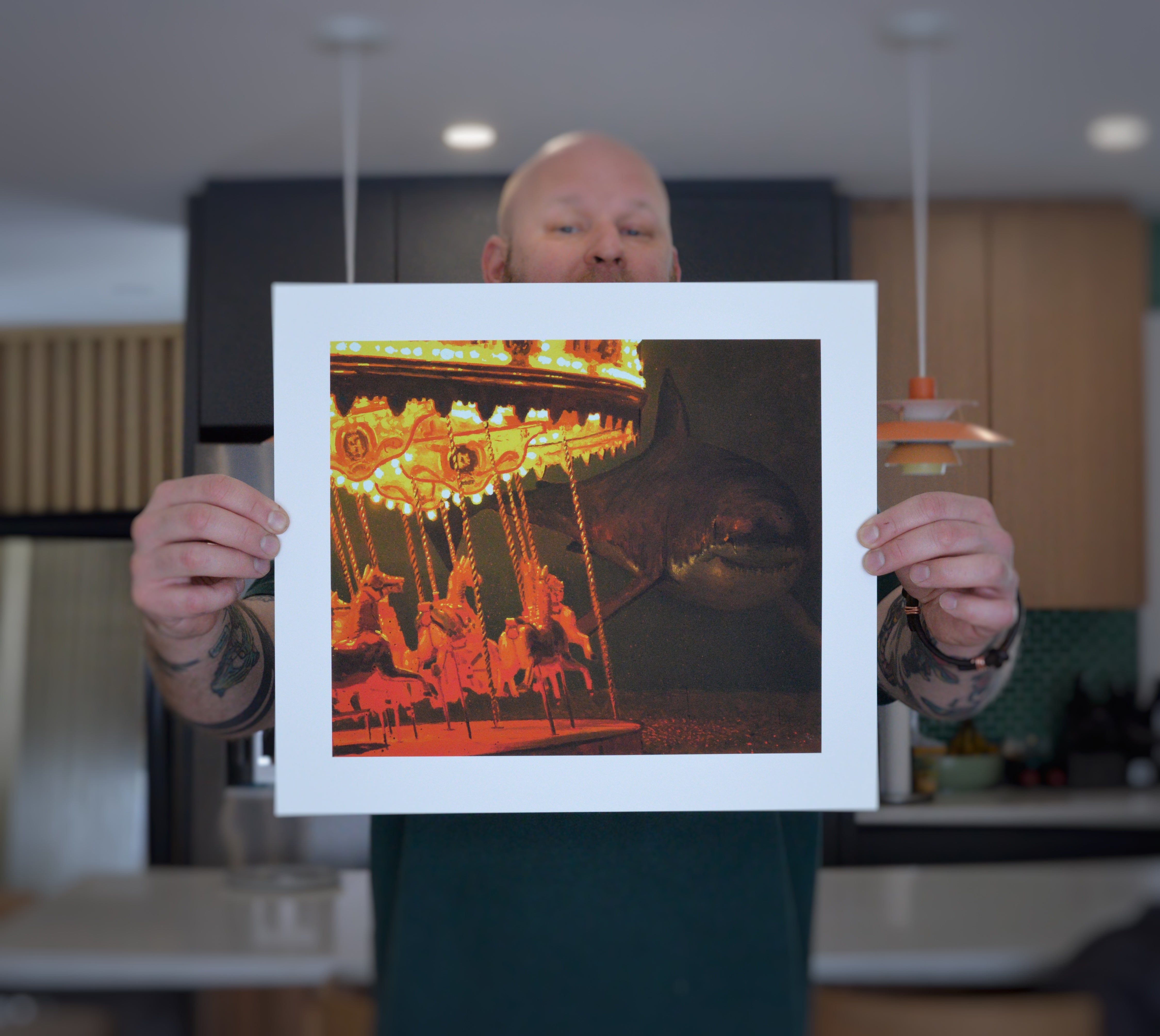Someone holds a photo titled Take Me To The Carnival by Chris Austin, showcasing a shark on a carousel amidst brightly lit, colorful horses, evoking a vibrant carnival. The background reveals a blurred kitchen interior with modern lights and furniture as an Archival Pigment Print.