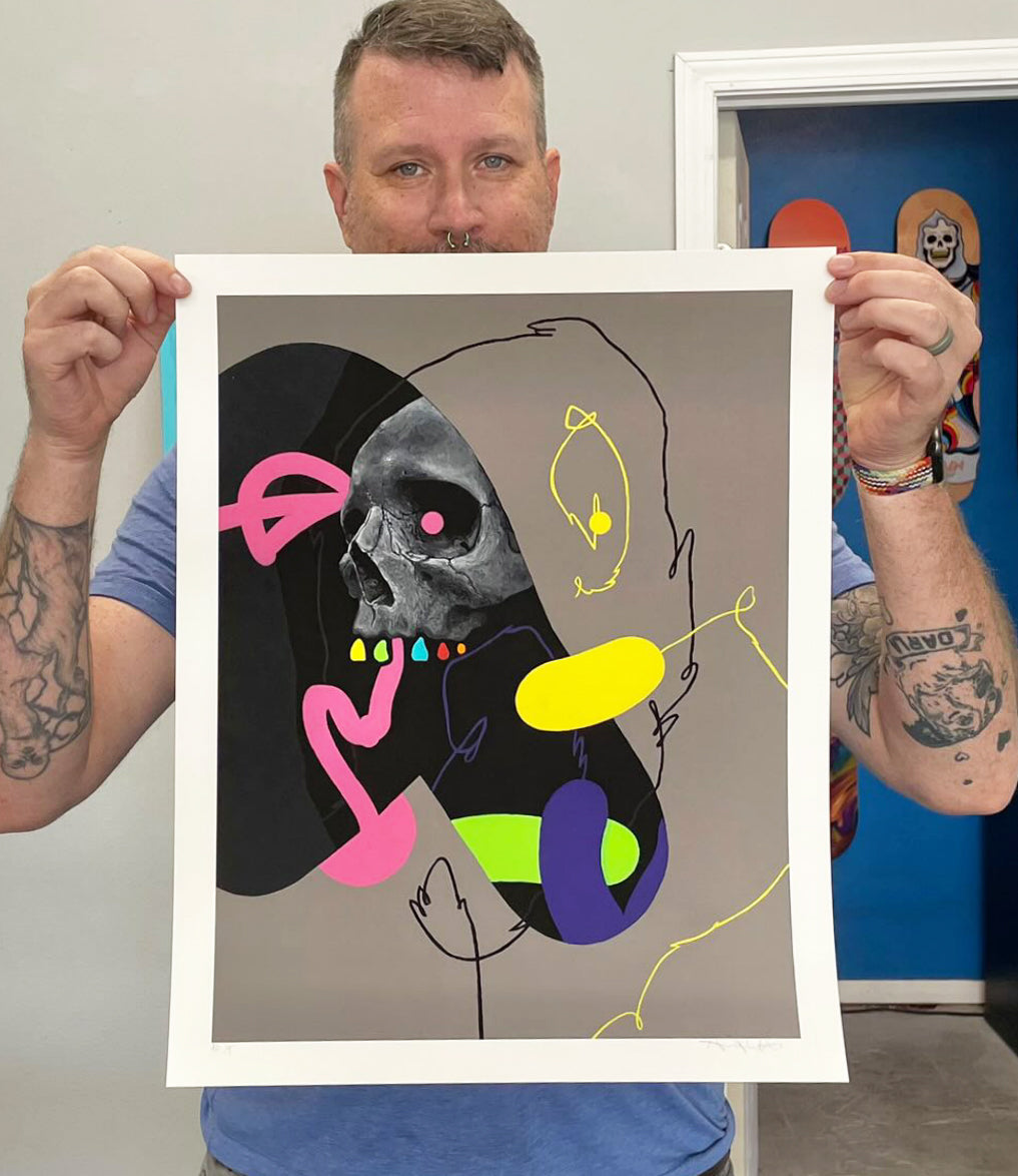 A tattooed individual displays Ive Seen This Side Of You, a signed, numbered piece by Anthony Hurd. It features an abstract skull blending pink, yellow, blue, and black shapes on gray. They stand indoors with a vibrant painting in the backdrop.