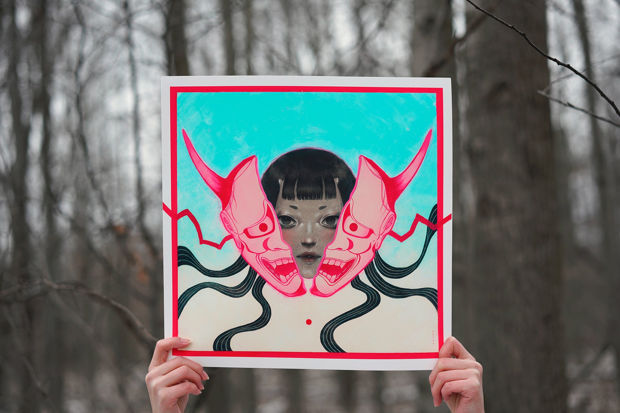 A person holds up Shoko Ishida’s “HANNYA,” an archival pigment print featuring two red demon masks and swirling black lines on blue, blending a real face with the artwork; bare trees appear in the background.