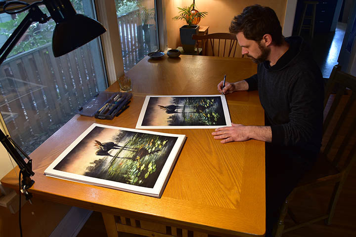 In a well-lit room with a view of greenery, someone signs two limited edition Islands prints by Brin Levinson, depicting a moose standing in water with lily pads at a wooden table.
