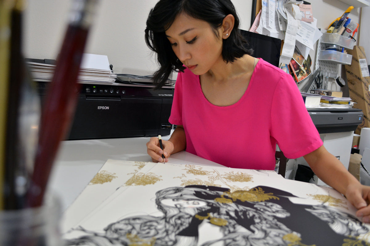 In a pink shirt, a woman carefully creates The Yellow Passerby & I by Tran Nguyen at her desk with art supplies and a printer, focusing on producing an intricate screenprint destined to be signed by the artist.