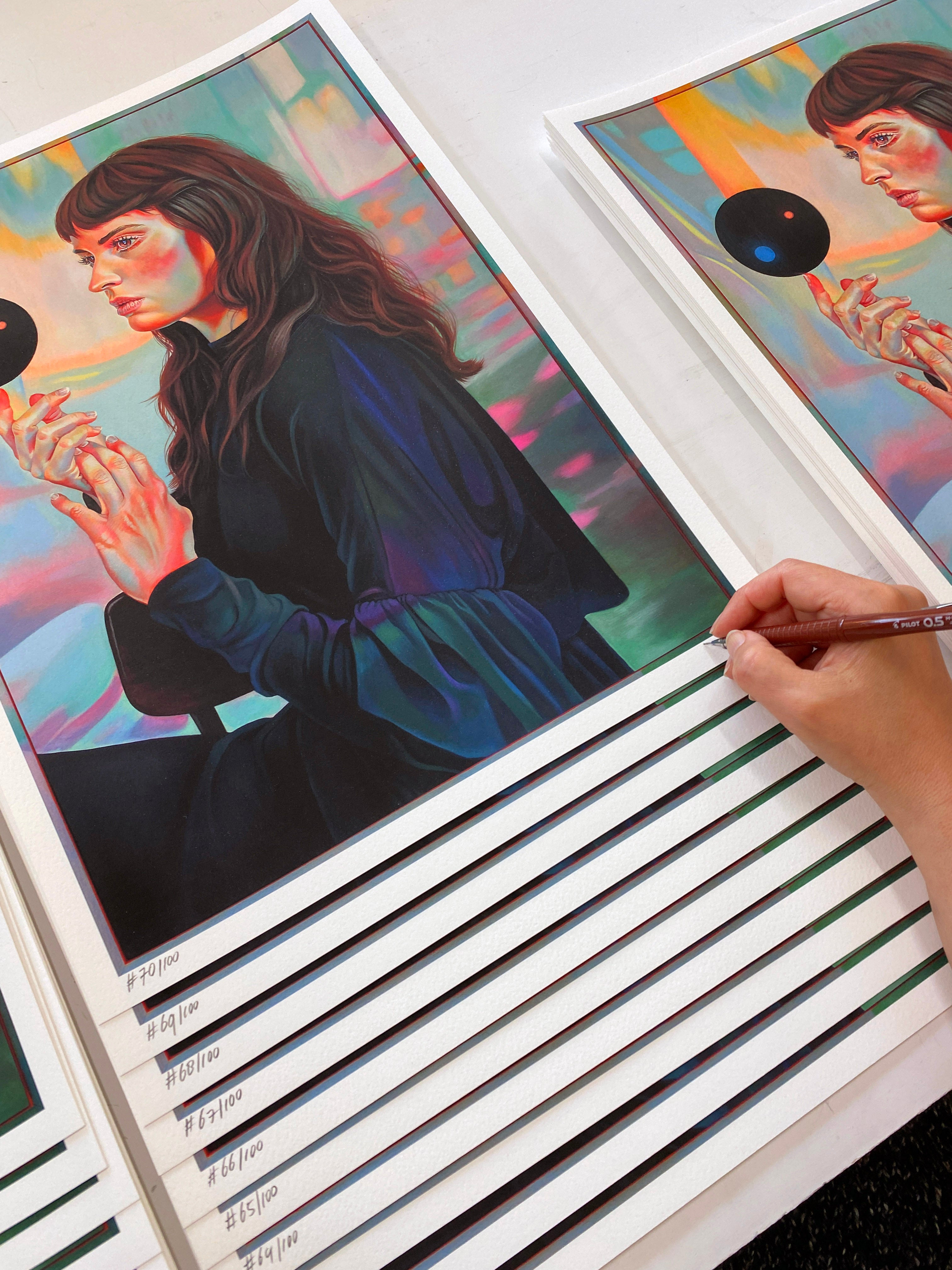 A person signs Power of Three by Martine Johanna, a series of vibrant archival pigment prints depicting a woman in a dark dress with a black circle. Each print is numbered, ready for signing, and artistically stacked.