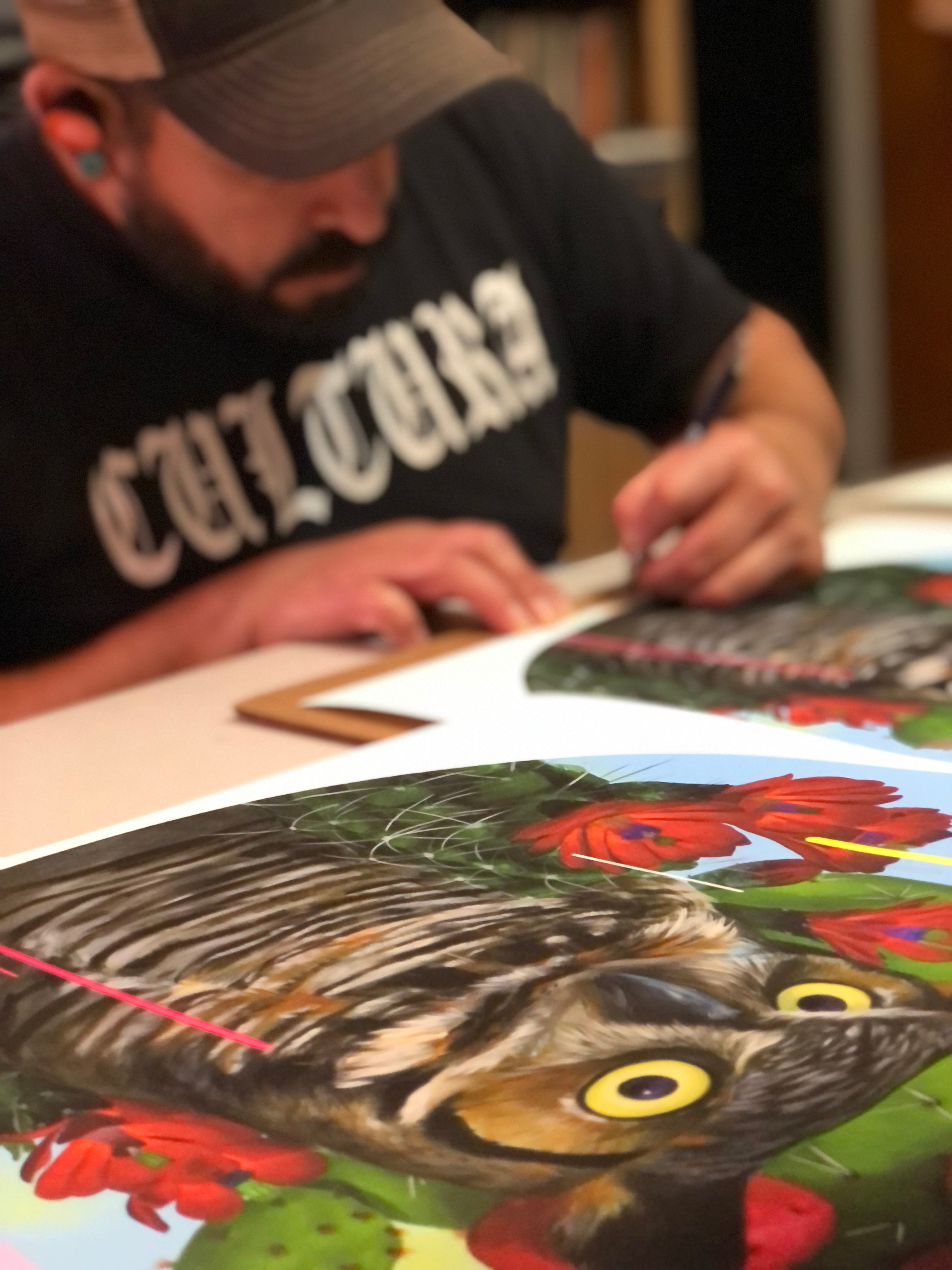 A man in a cap and a black Cultura shirt focuses on signing an archival pigment print titled Santuario by Frank Gonzales, featuring a vibrant tiger among red flowers. The striking artwork dominates the scene, with his partially blurred face in the background.