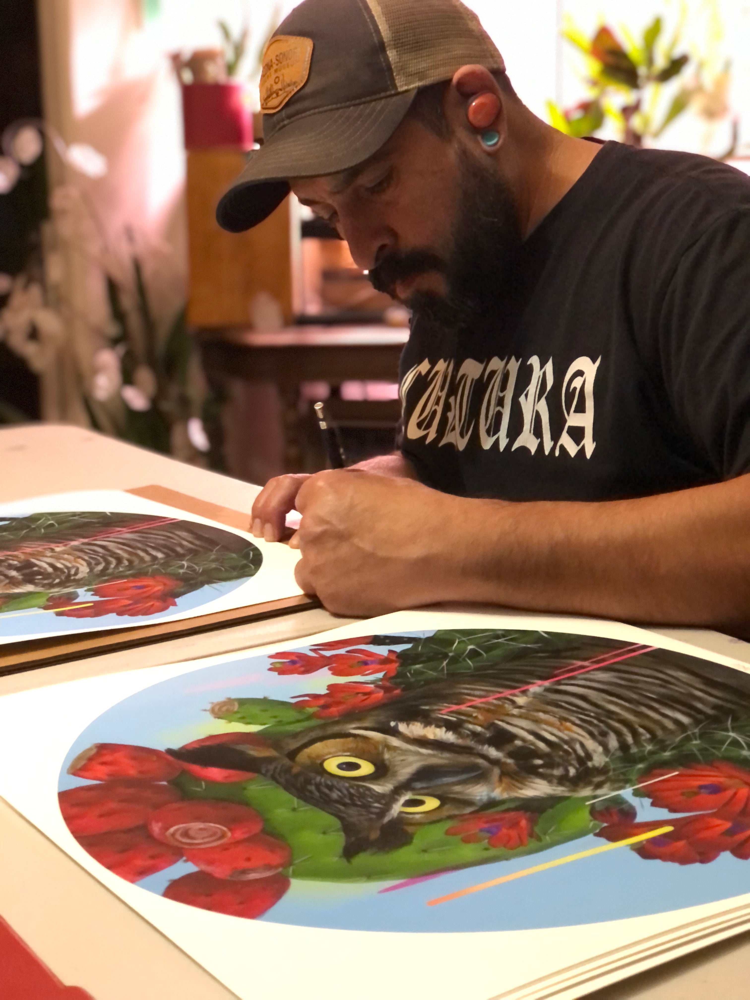 In an art studio, a bearded man wearing a cap and T-shirt creates Santuario, an archival pigment print by Frank Gonzales. This circular piece depicts an owl with vibrant eyes surrounded by red flowers, plants in the background, and is part of a signed and numbered edition.