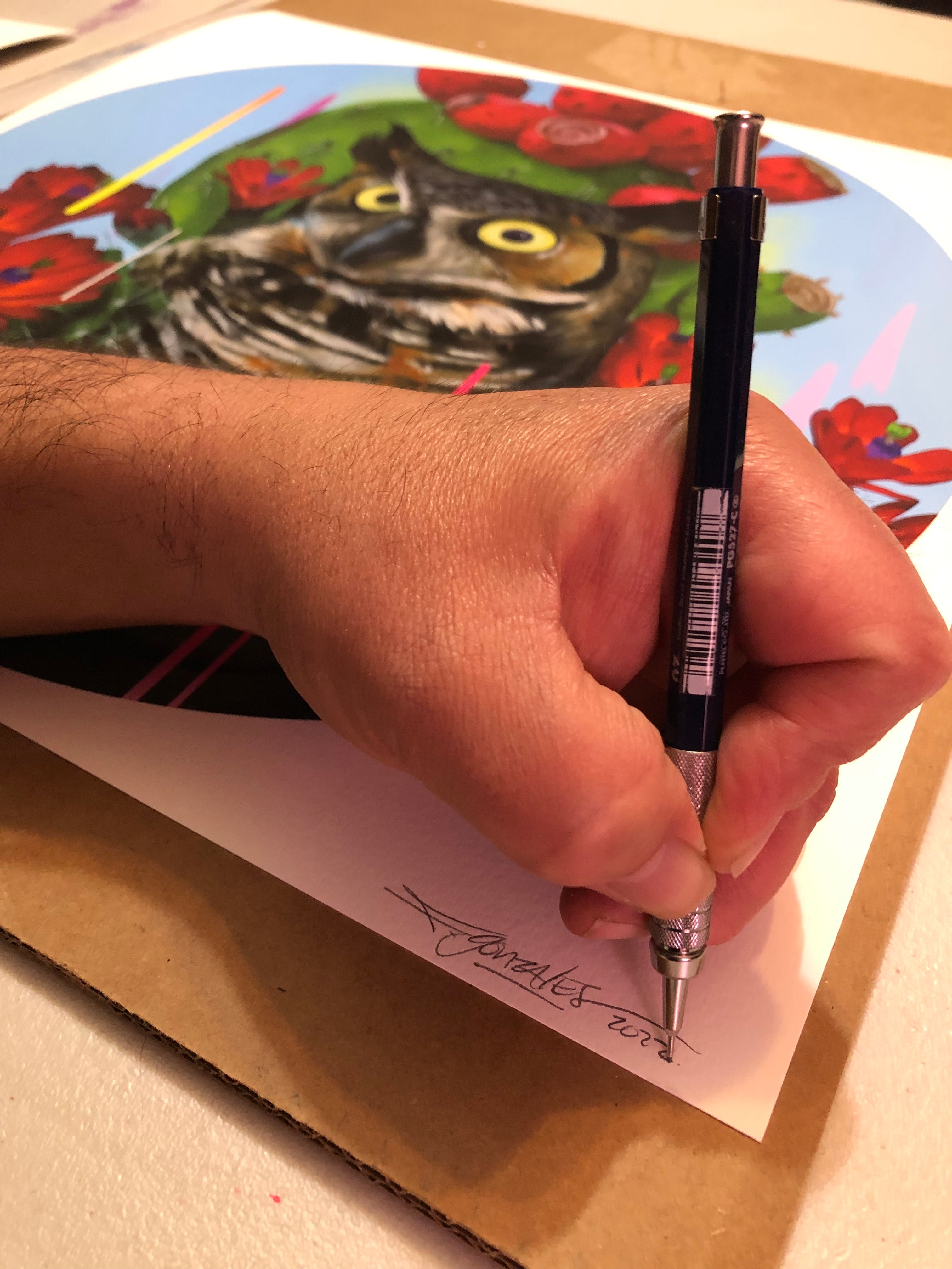 A hand signs Santuario, an archival pigment print by Frank Gonzales, depicting a colorful owl amid red flowers. The signer’s arm is partially visible on the artwork, part of a signed and numbered edition on cardboard.