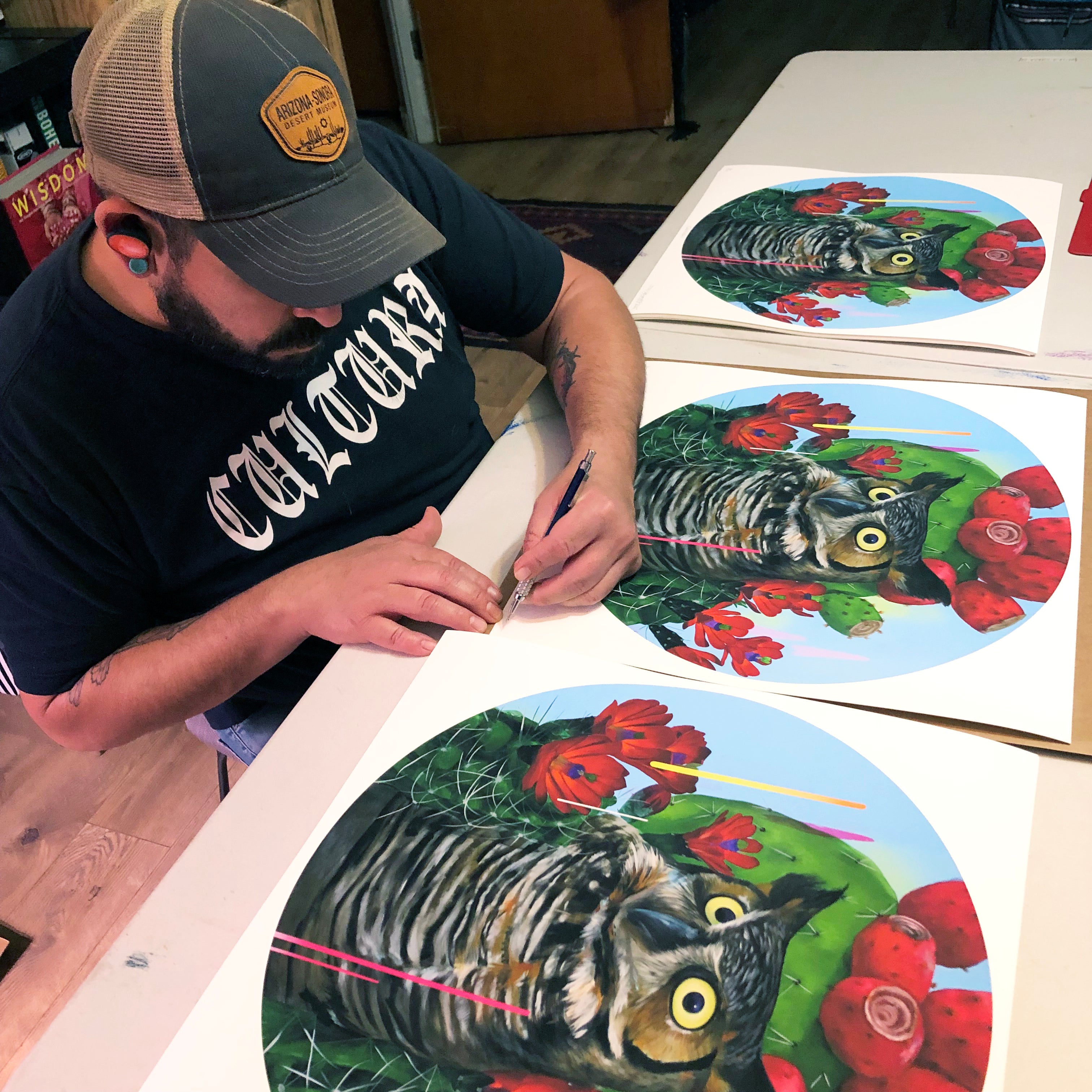 A person in a black T-shirt is focused on drawing on an Santuario archival pigment print by Frank Gonzales, which features an owl surrounded by red flowers and cacti. The signed, numbered edition rests among similar prints on the table, capturing their artistic attention.