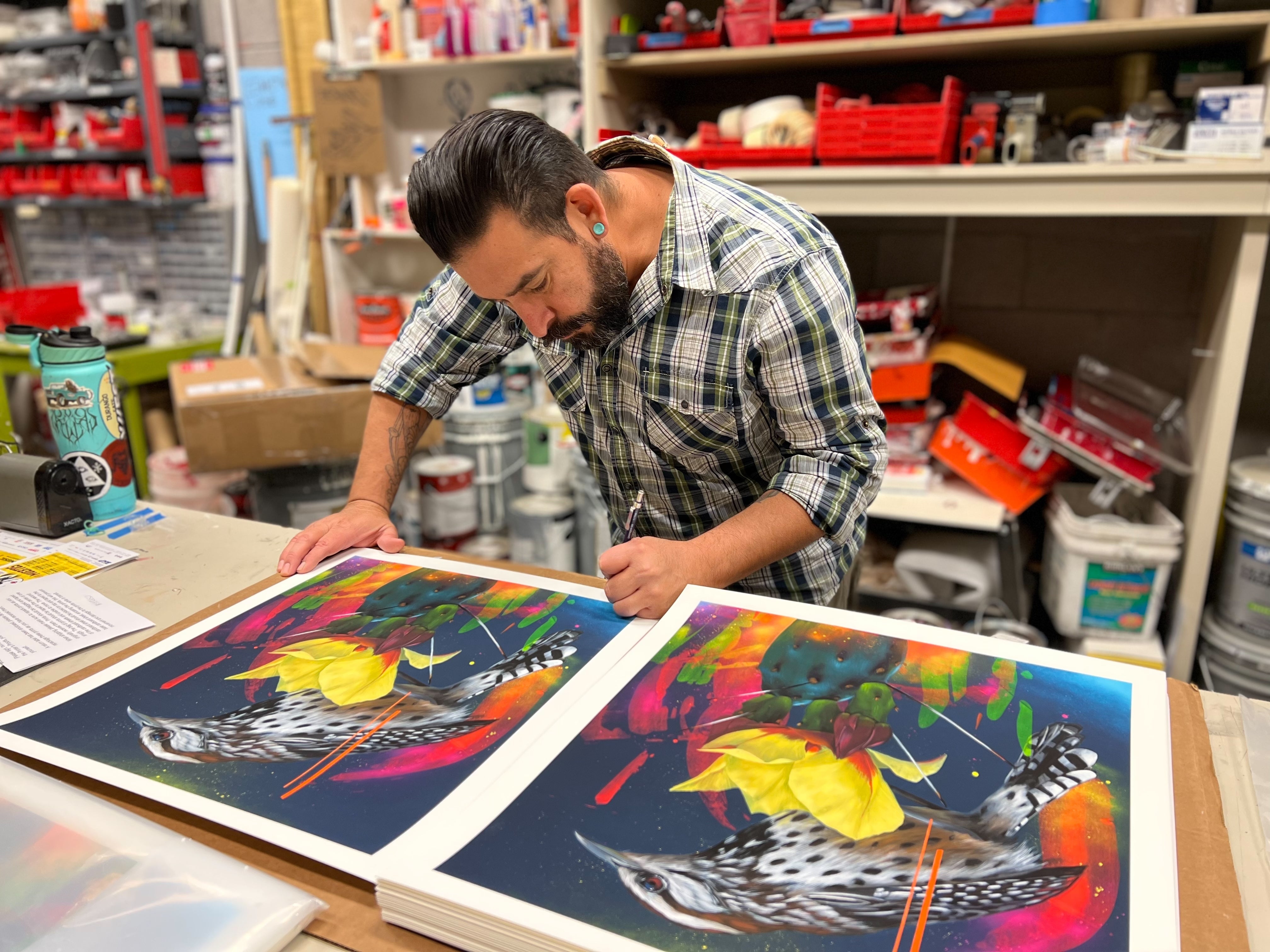 Frank Gonzales signs and numbers "Monsoon Misterioso" archival pigment prints, featuring a bird, leaves, and abstract shapes at his studio worktable surrounded by art supplies and tools.