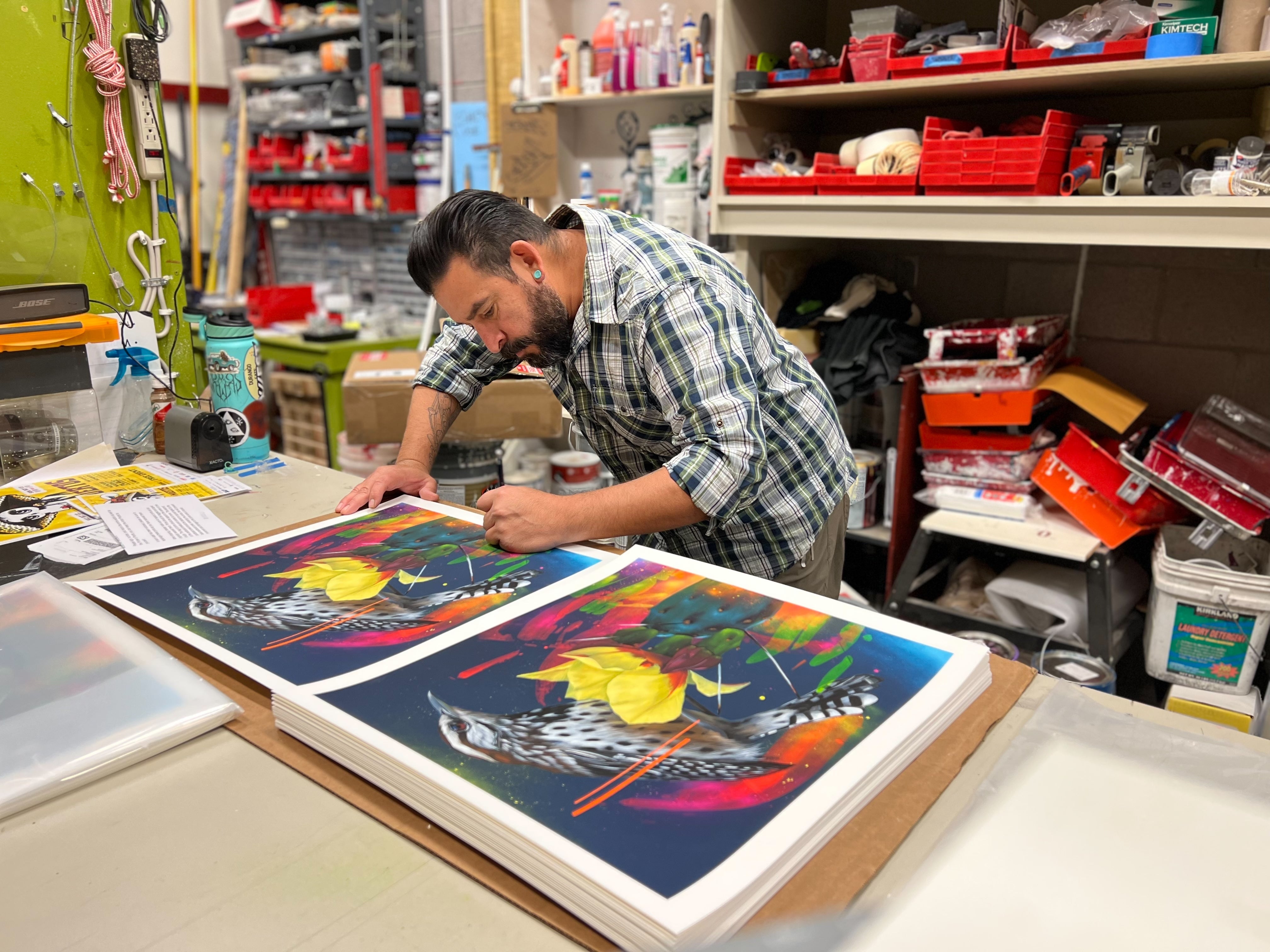 In his art studio, Frank Gonzales signs and numbers "Monsoon Misterioso" archival pigment prints, featuring a fish and leaves, surrounded by tools, papers, and art supplies.