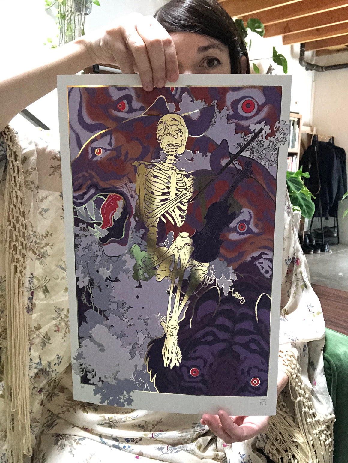 Someone proudly displays Soothe by JAW Cooper, featuring a skeleton with a smoking pipe against a vibrant backdrop. Abstract shapes and eyes are part of the design, while the persons patterned clothing partially hides their face, enhancing the artworks charm.