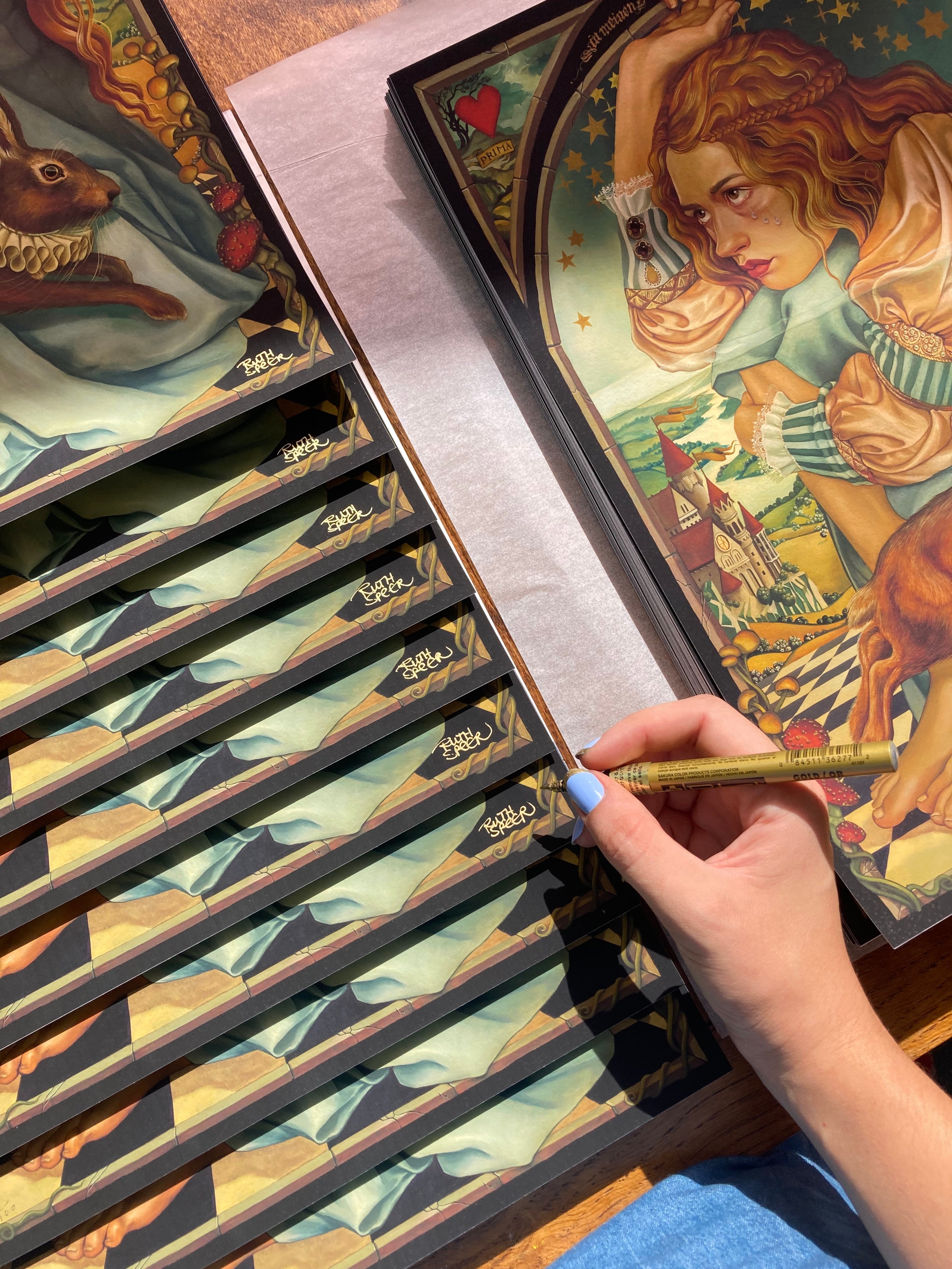 A person with light blue nail polish signs a Ruth Speer poster titled Alice. It features a surreal scene with a woman, rabbit, and whimsical elements. Hand-embellished posters are signed, numbered, and stacked on the left.